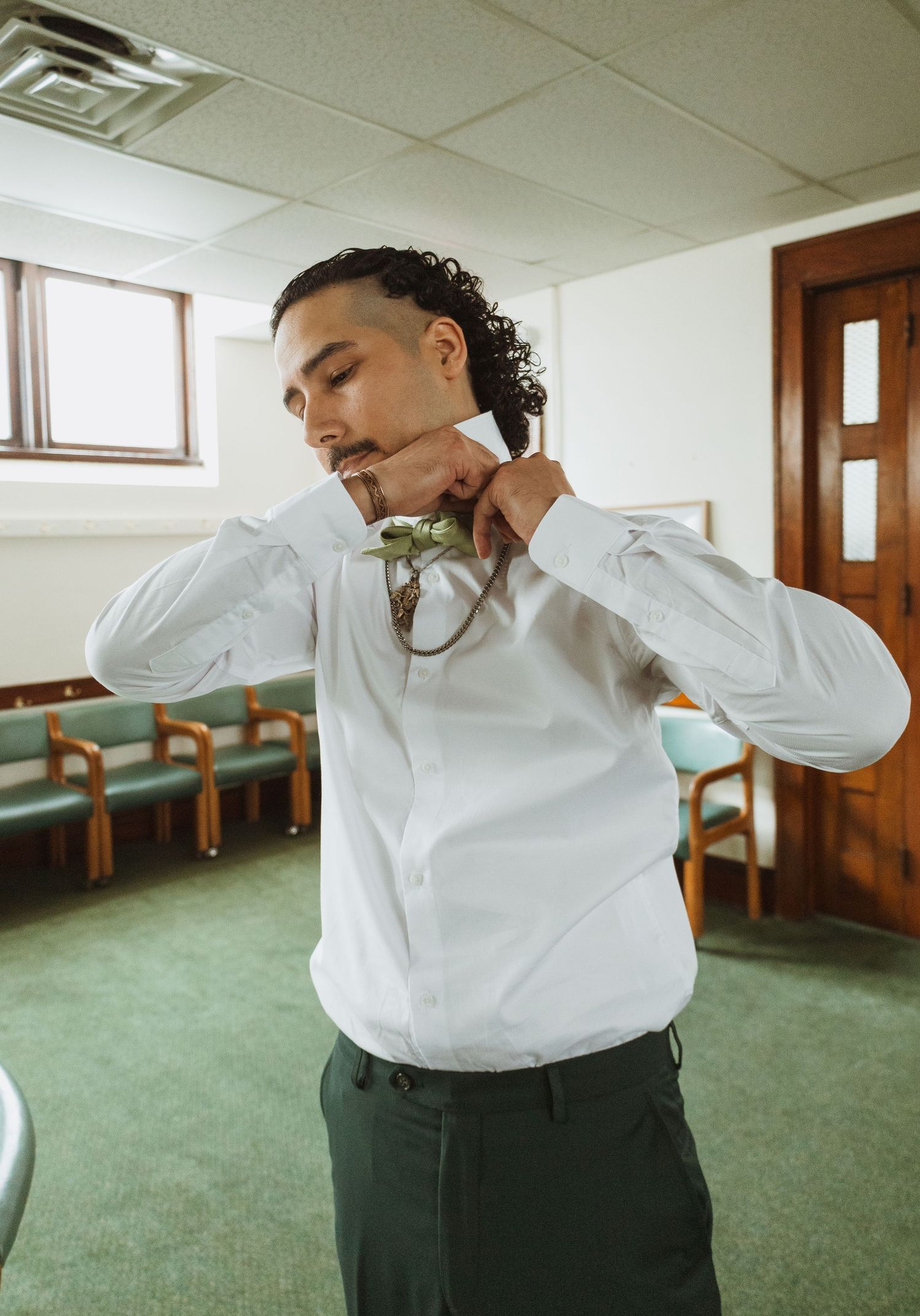 Groom in white shirt and dark pants tying green bow tie in a room with chairs.