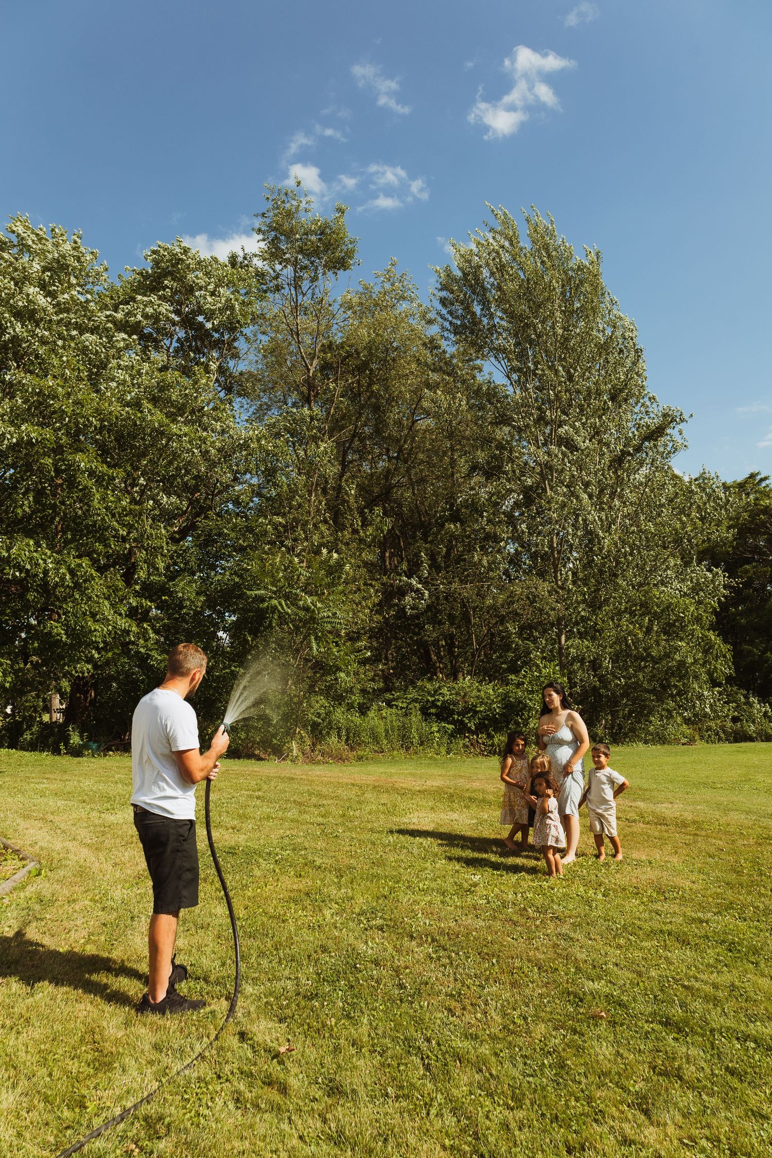 Family enjoying summer day in garden, man spraying water with hose while woman and children play on green lawn.