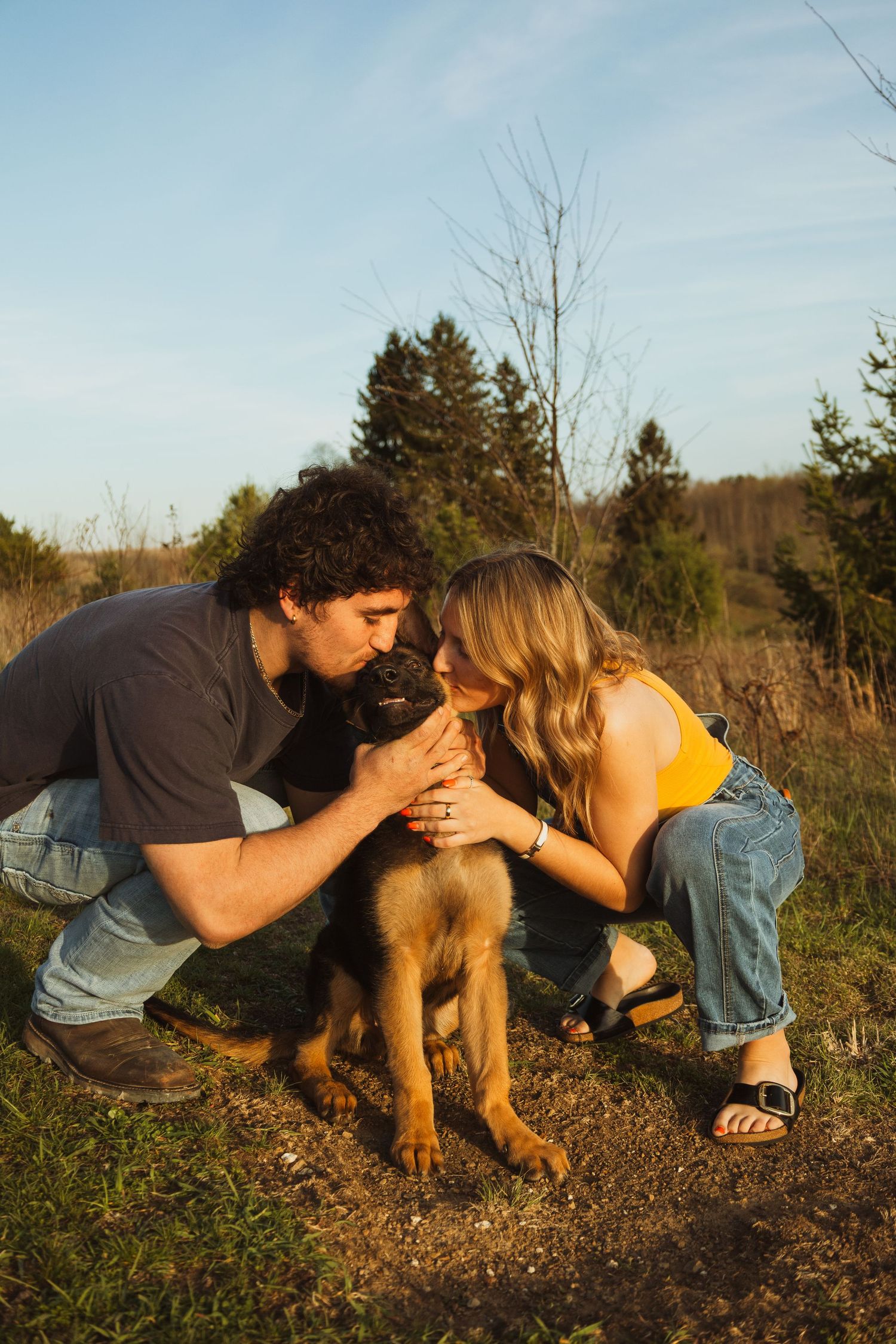 A couple crouches outdoors kissing their brown puppy at golden hour with trees and blue sky in the background.