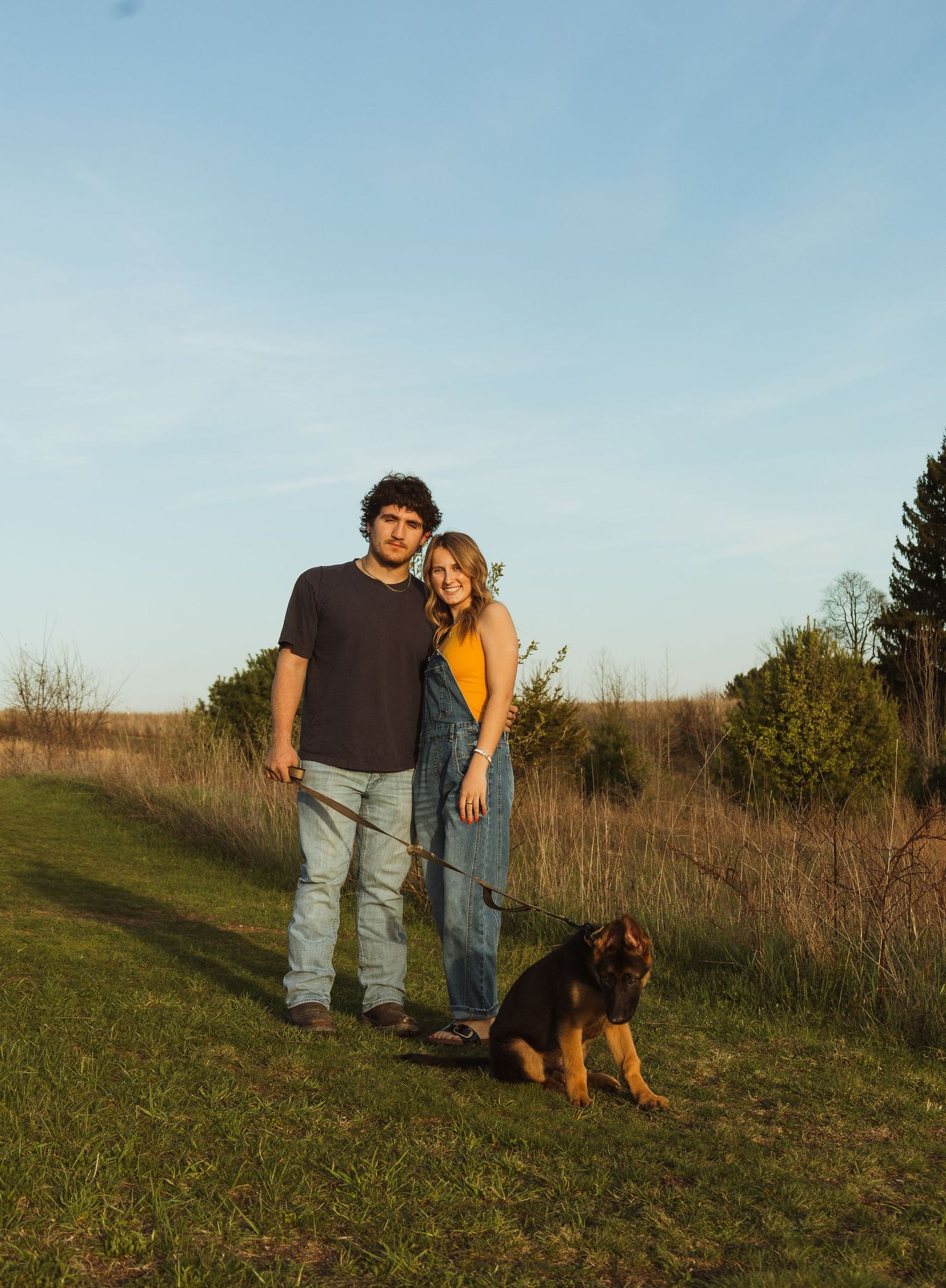 A young couple stands smiling on a grassy path at sunset with their German Shepherd dog beside them.