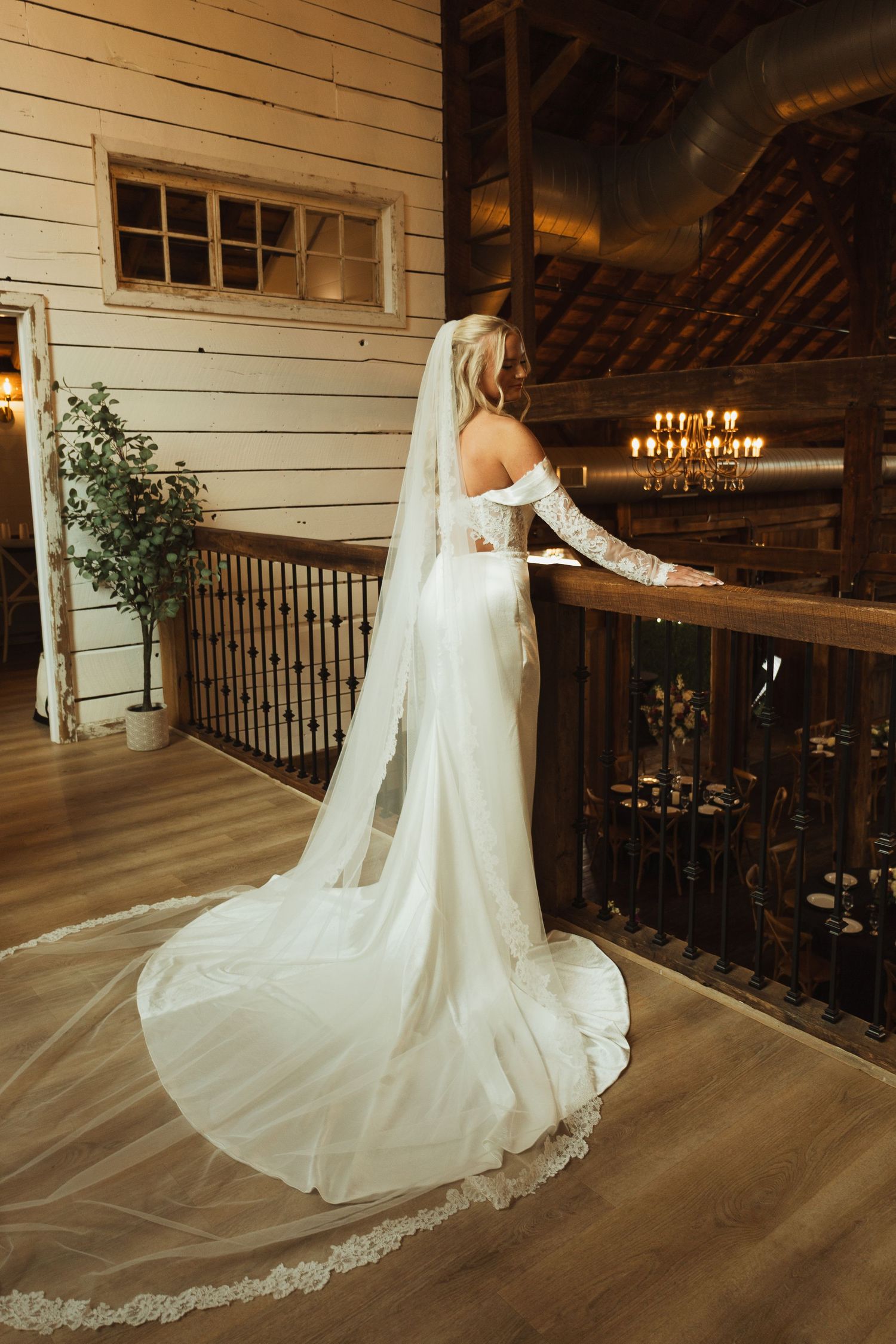 Bride in elegant off-shoulder white gown with lace-trimmed cathedral veil stands on rustic barn venue balcony.
