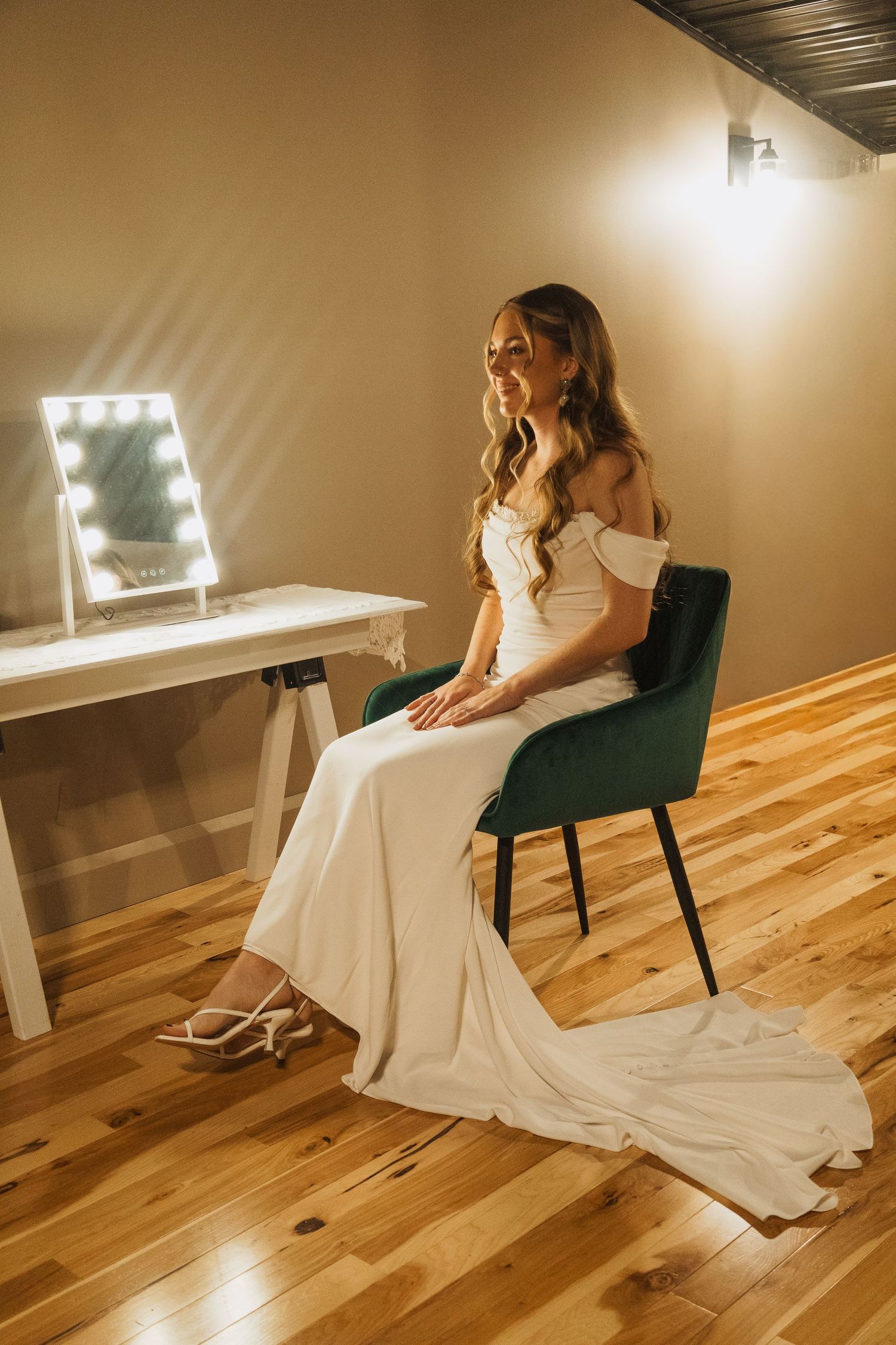 A bride in an elegant white gown sits at a vanity with illuminated mirror in a preparation room.