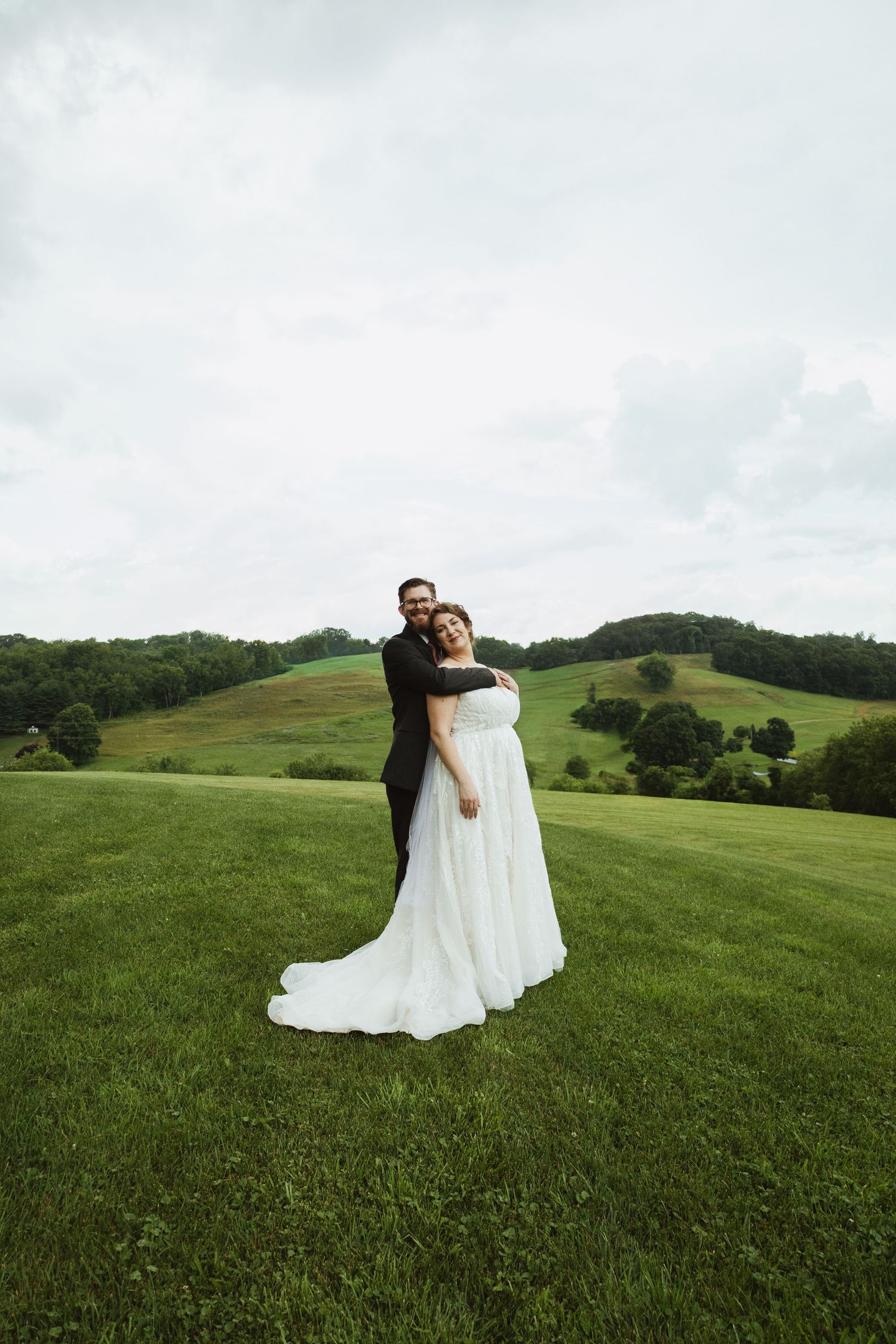 A couple embraces on a lush green hilltop meadow with rolling hills in the background on their wedding day.