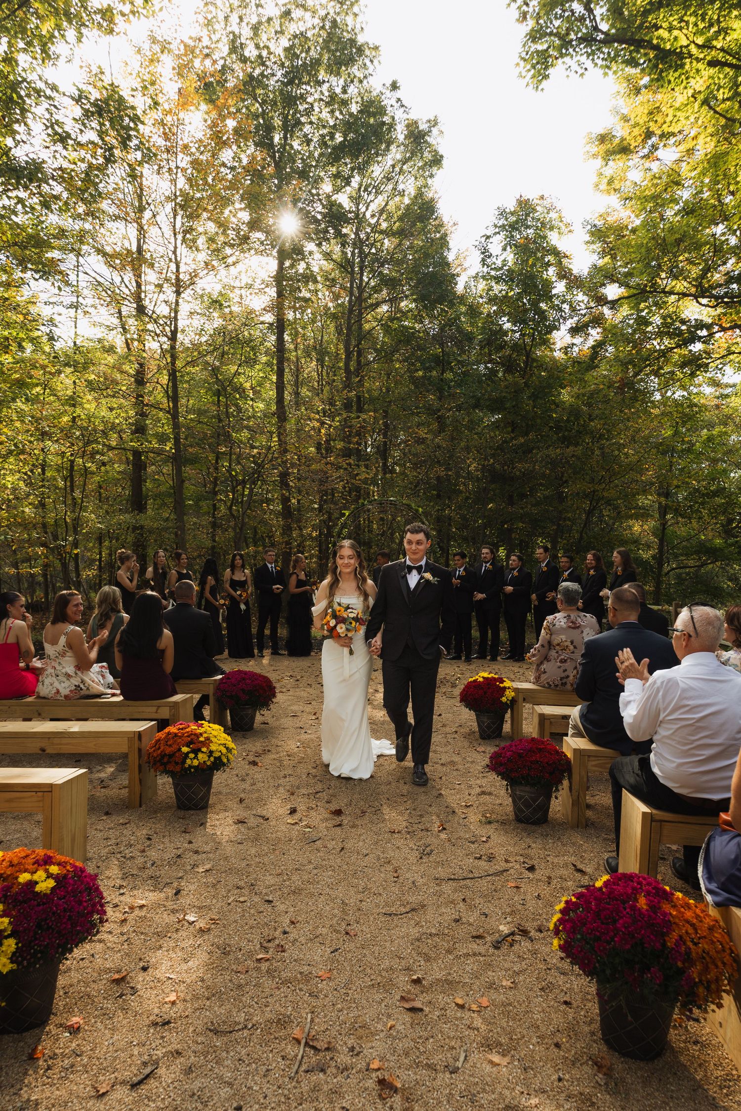 Outdoor wedding ceremony in a wooded area with red mums and wooden benches.