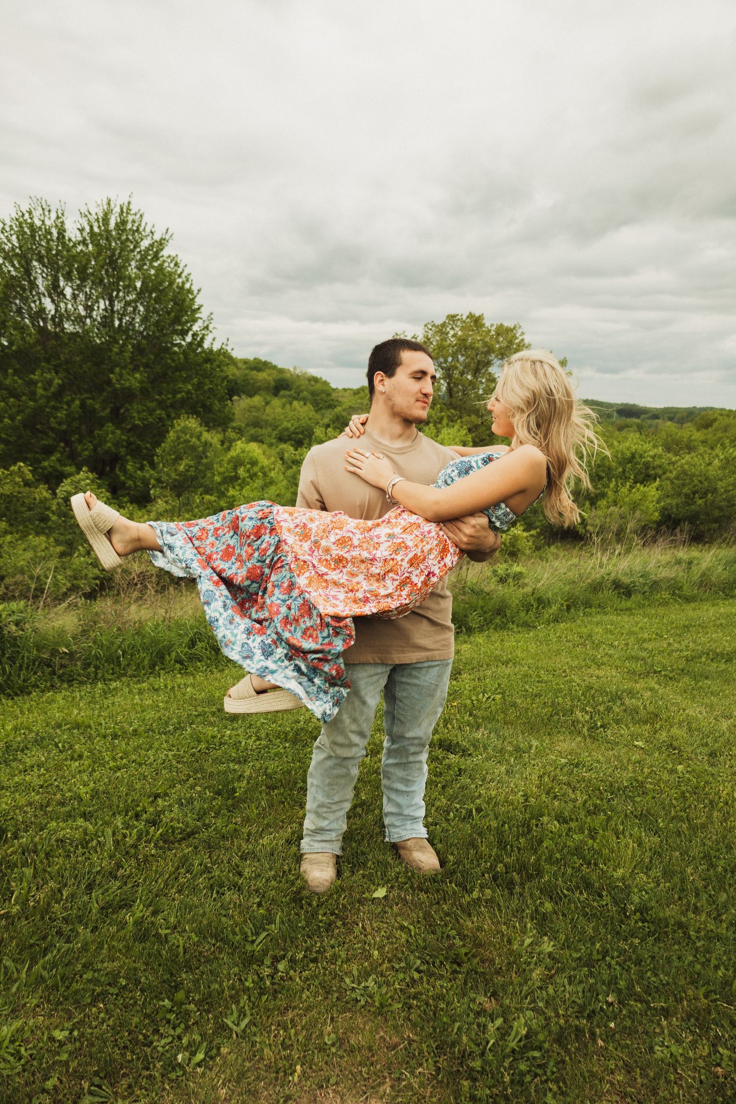 A couple shares a romantic moment in a grassy field under cloudy skies as he holds her in his arms.