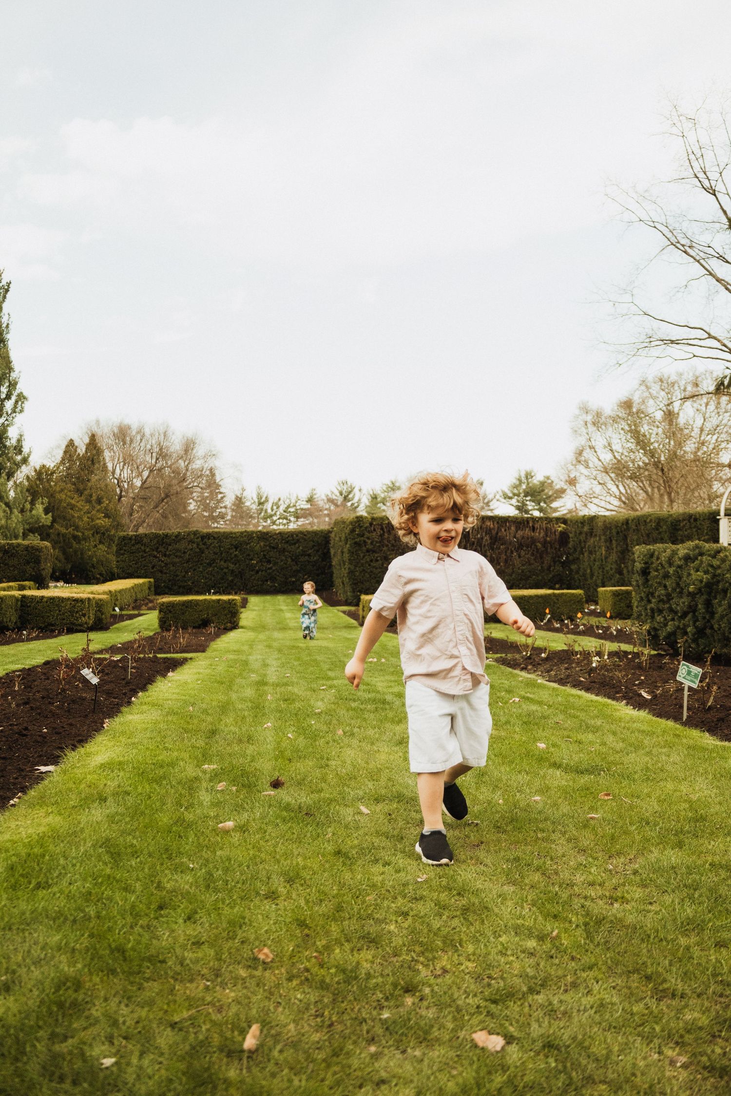 A child runs and plays in a well-manicured garden with tall hedges and grass paths on a cloudy day.