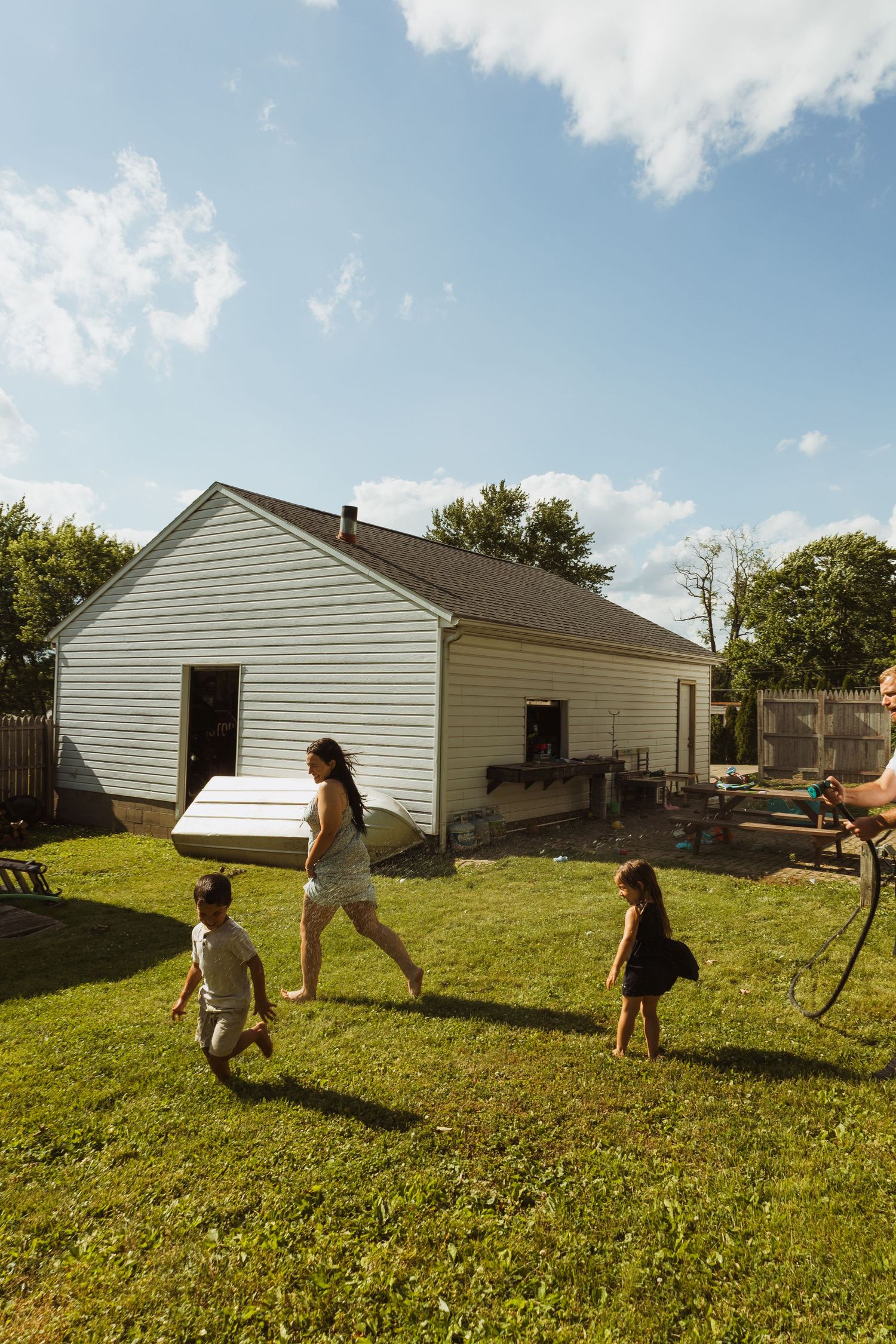 Family carrying items across a sunny backyard with a white house, green lawn, and blue sky on a summer day.