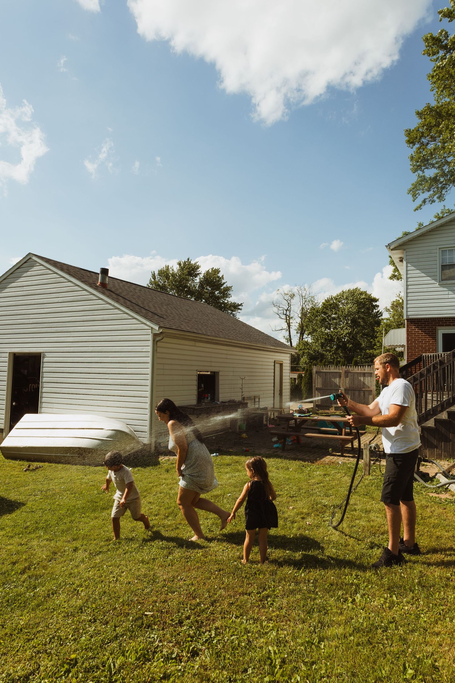 A man sprays water from a hose at two children and a woman running across a sunny backyard beside a white house.