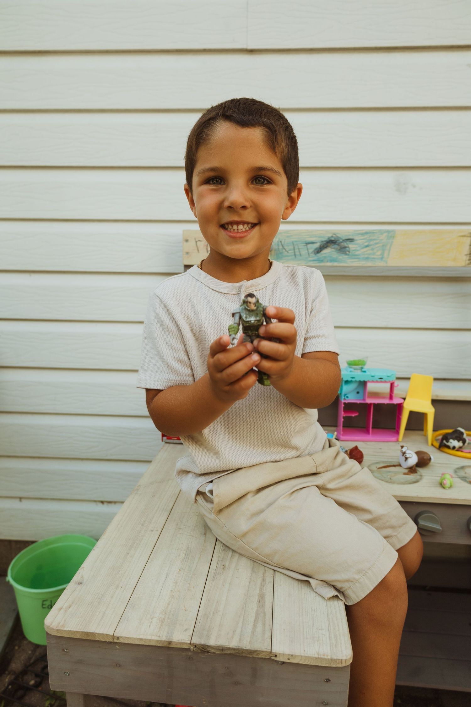 Smiling young boy sitting outdoors on wooden bench holding a toy action figure, wearing white t-shirt and beige shorts.