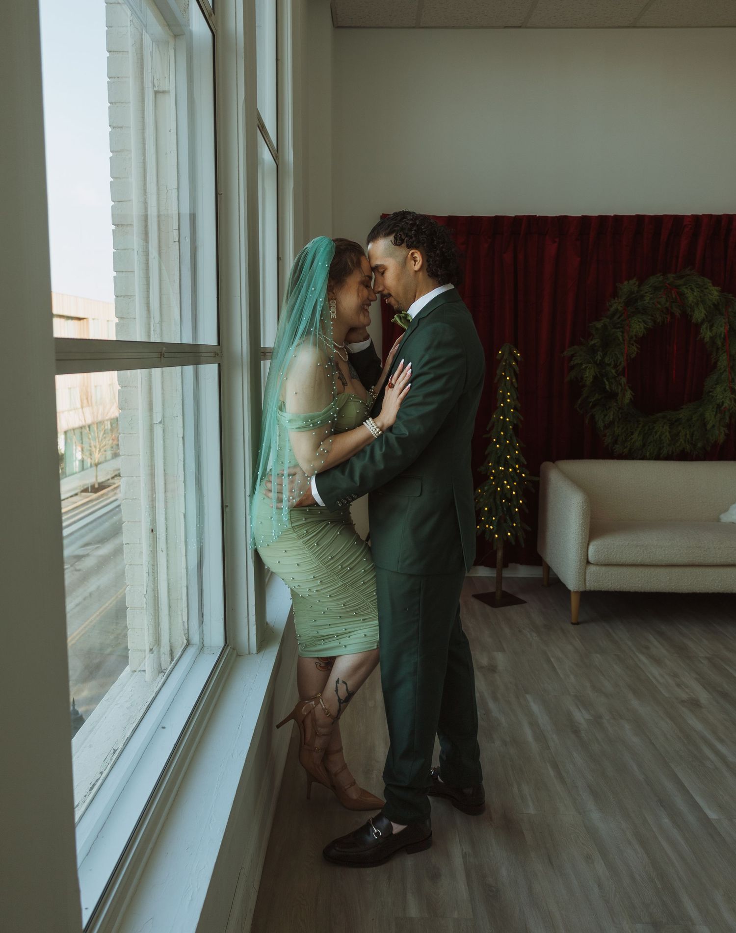 Bride in teal lace dress and green-veiled groom sharing intimate moment by large sunlit windows.
