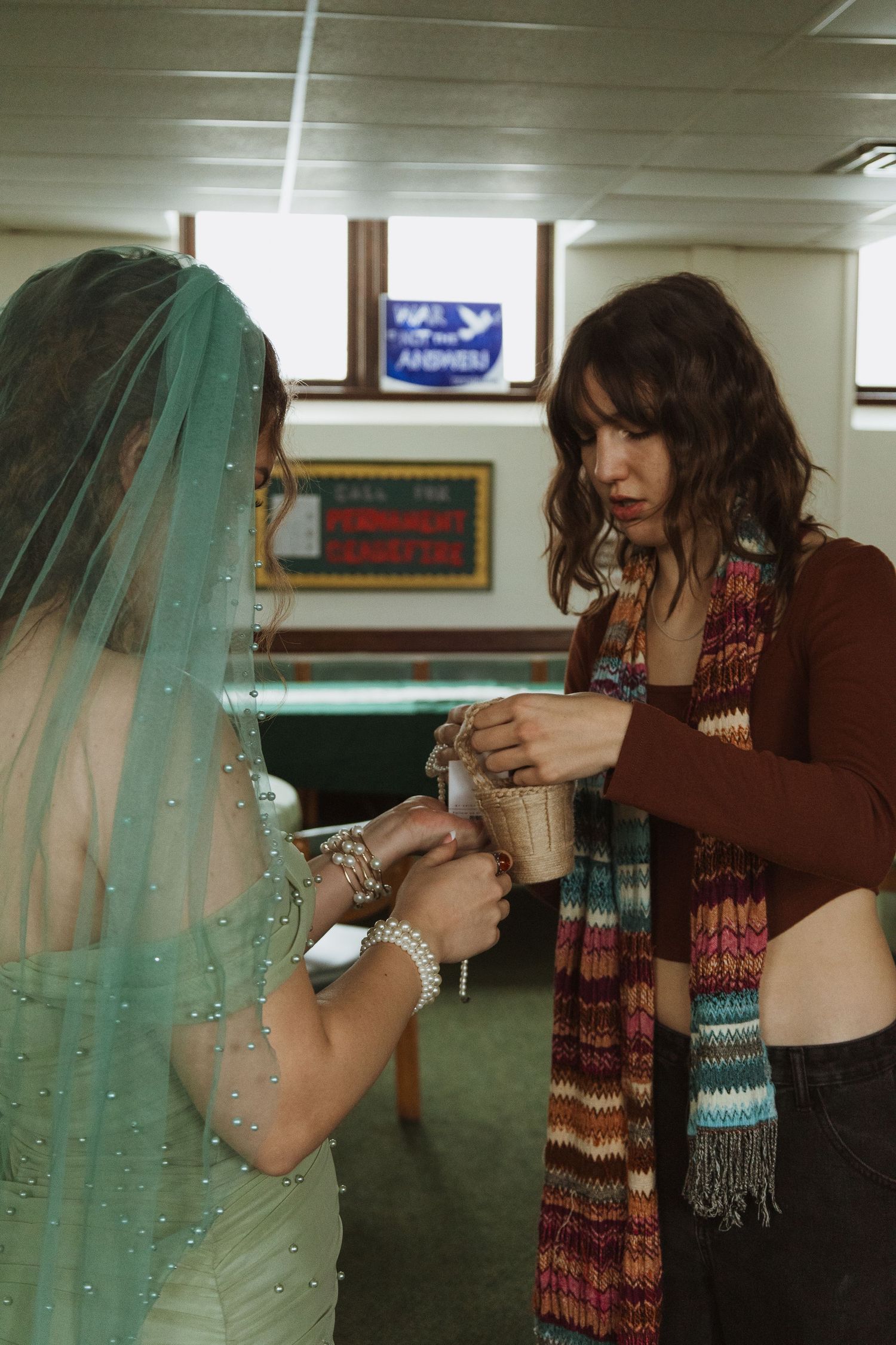 Bride in mint green beaded dress and teal veil receives help from bridesmaid wearing colorful scarf indoors.
