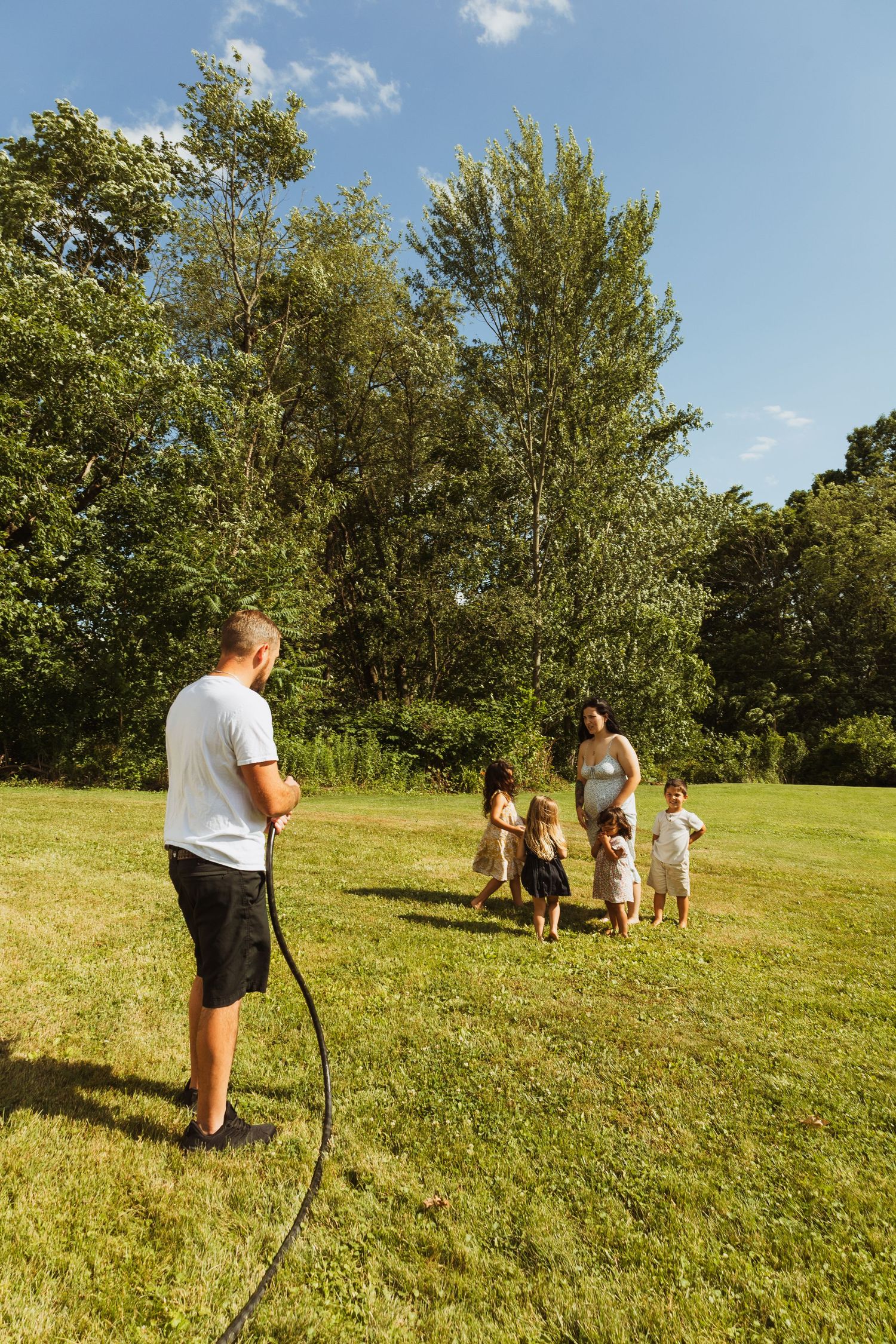 Family playing with a garden hose on a sunny green lawn surrounded by tall trees on a bright summer day.