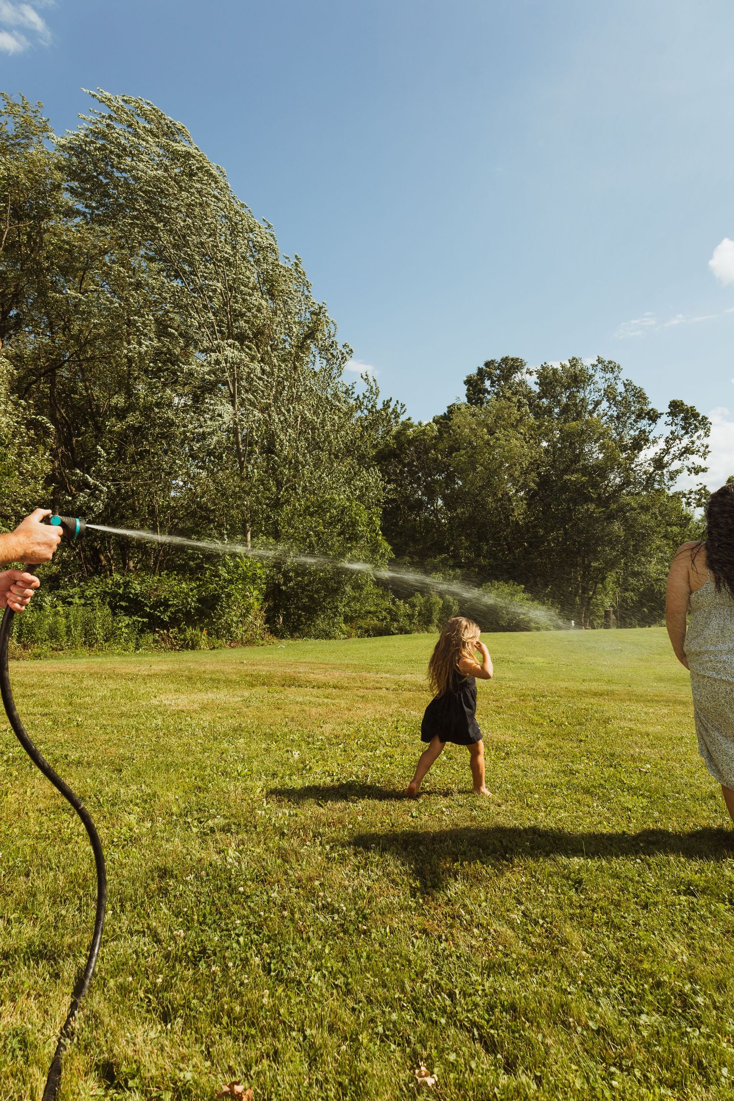 Children playing in a backyard with a garden hose on a sunny summer day, with green trees in the background.