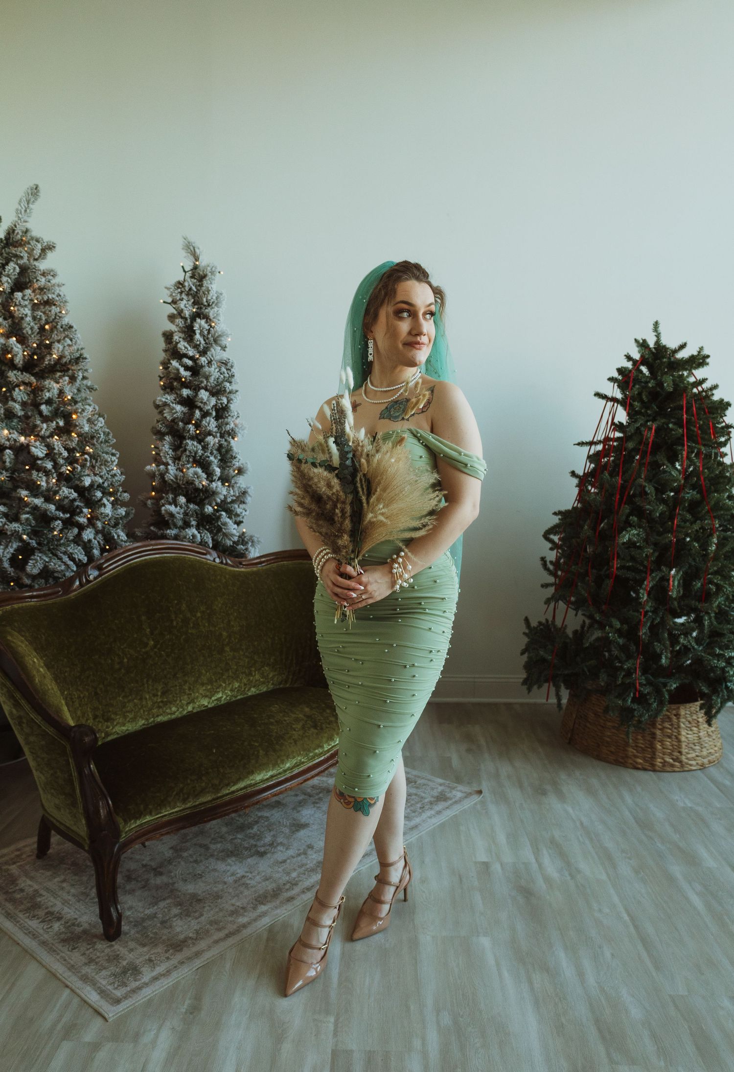Bride in sage green dress and teal veil posing beside Christmas trees indoors.