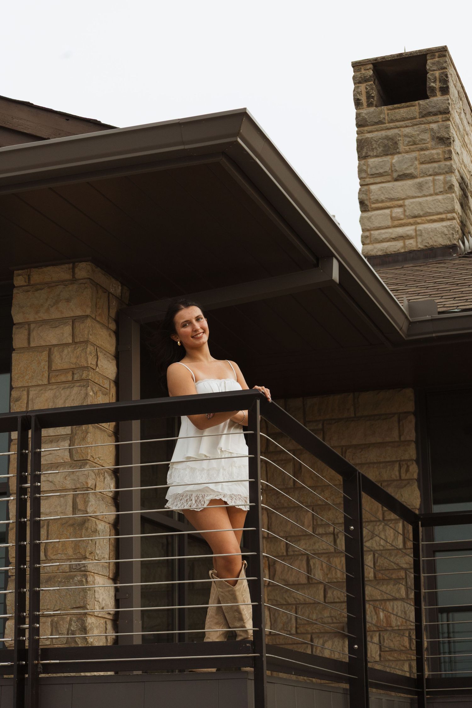 Young woman in white dress and boots smiling on modern stone home balcony with cable railing.