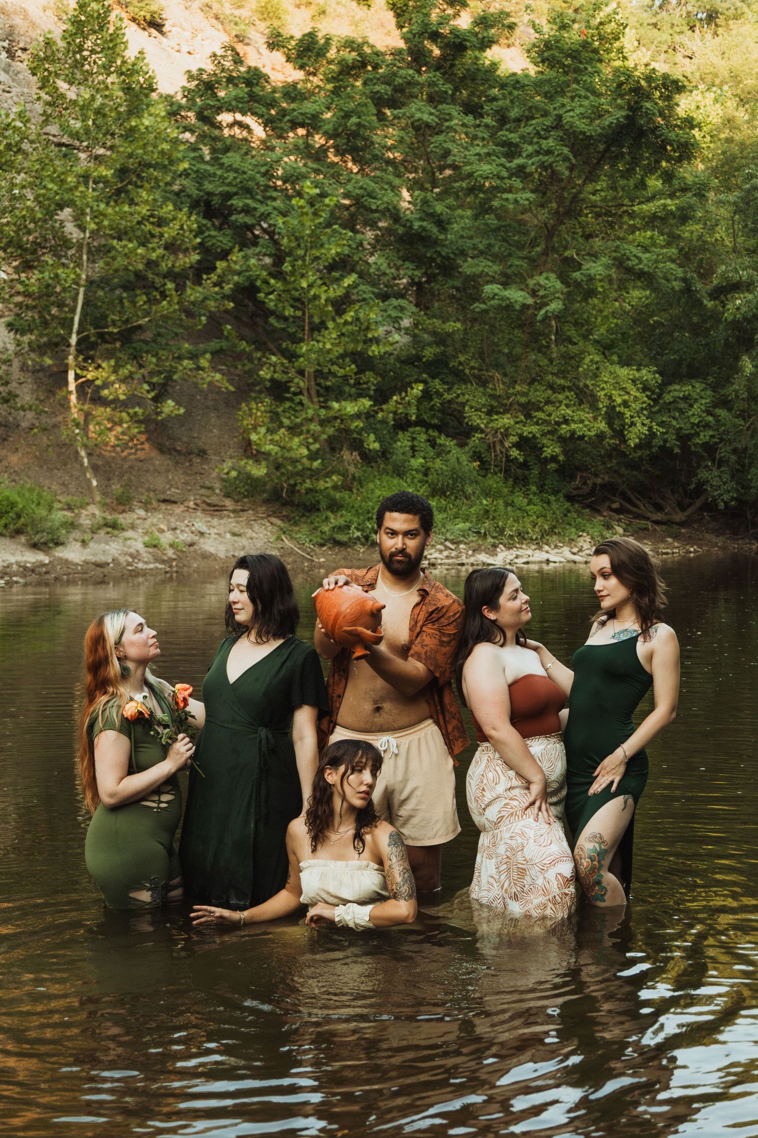 Group of six people standing in a river surrounded by lush green trees, man holding a pumpkin in center.