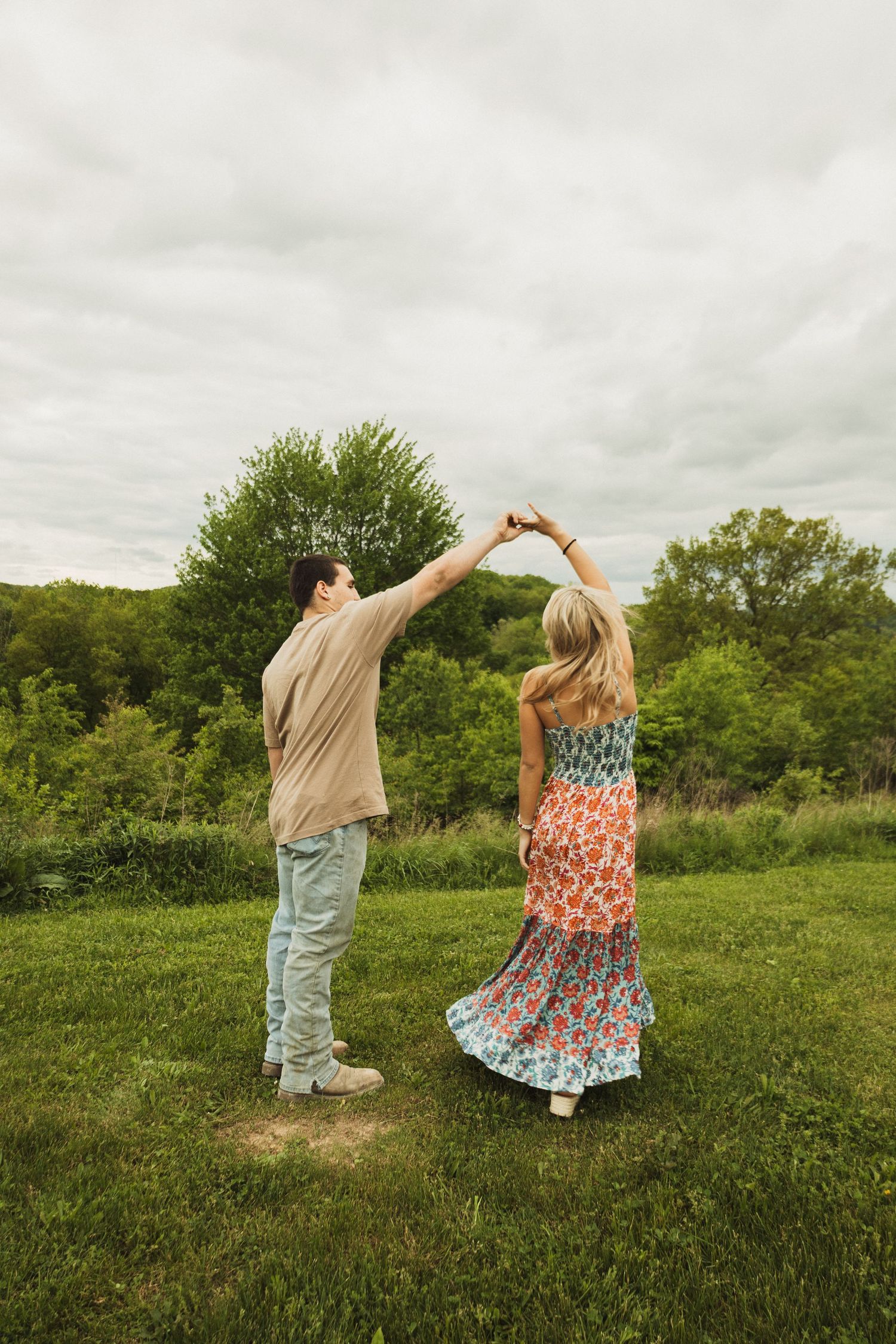 Couple dancing together on a hilltop, making a heart shape with their arms against a nature backdrop.
