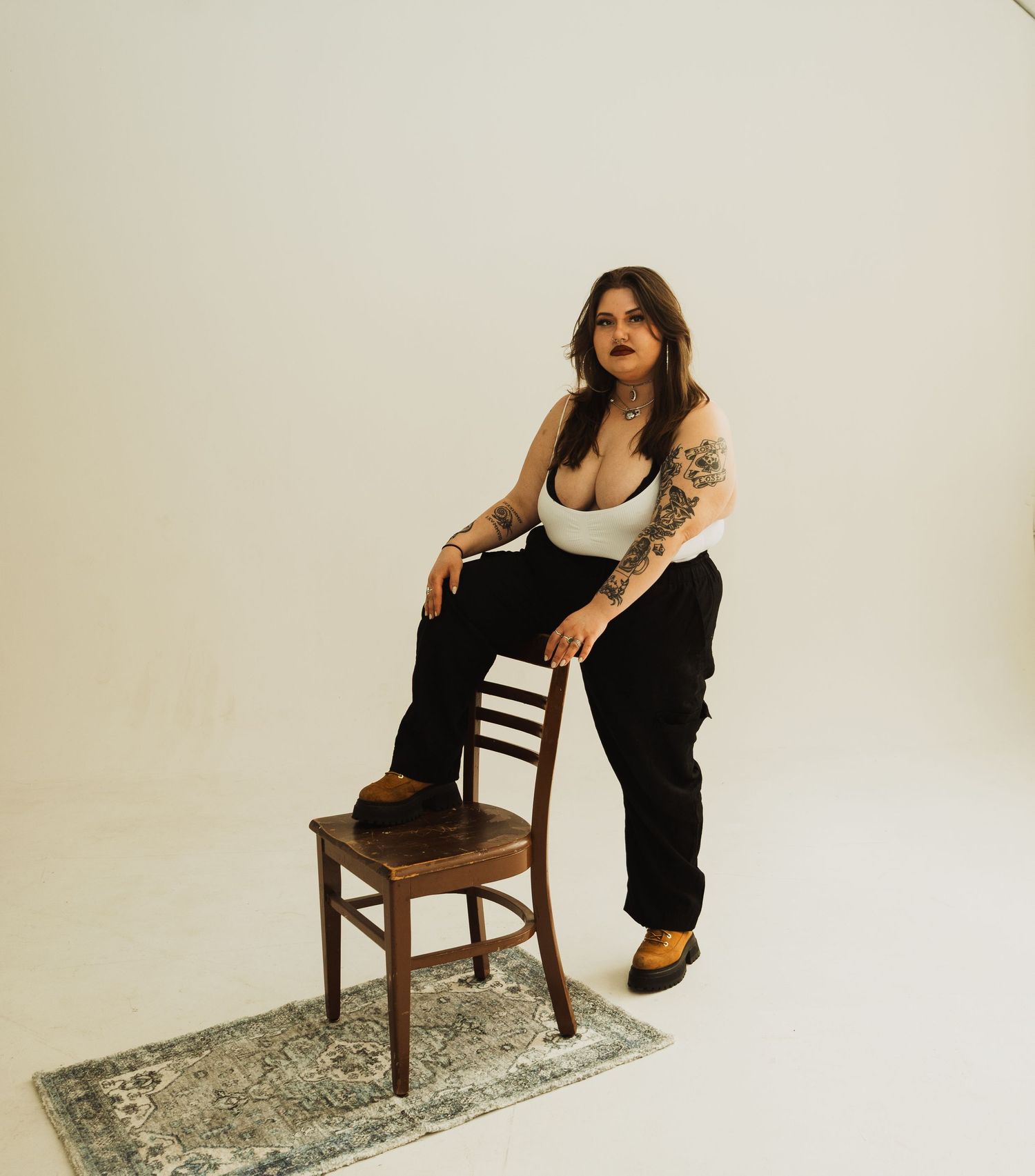 Model in white tank top and black pants poses against beige backdrop while leaning on vintage wooden chair.