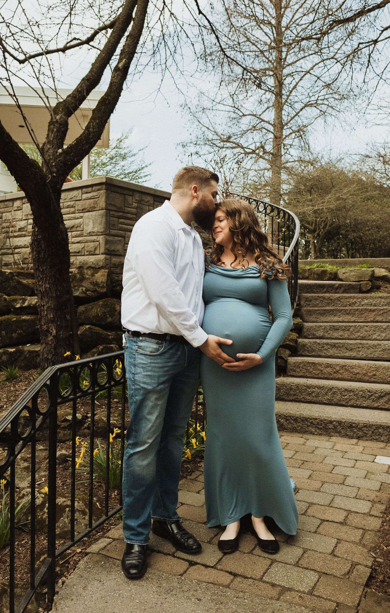 A loving couple embraces on stone steps as they celebrate their pregnancy in a teal maternity dress and white shirt.