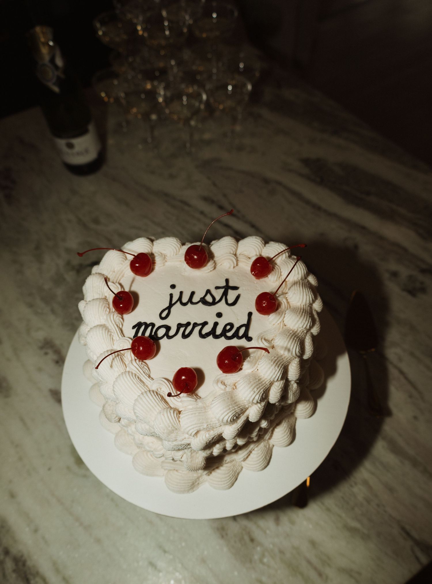 Heart-shaped white wedding cake decorated with red cherries and 'just married' text on marble surface.