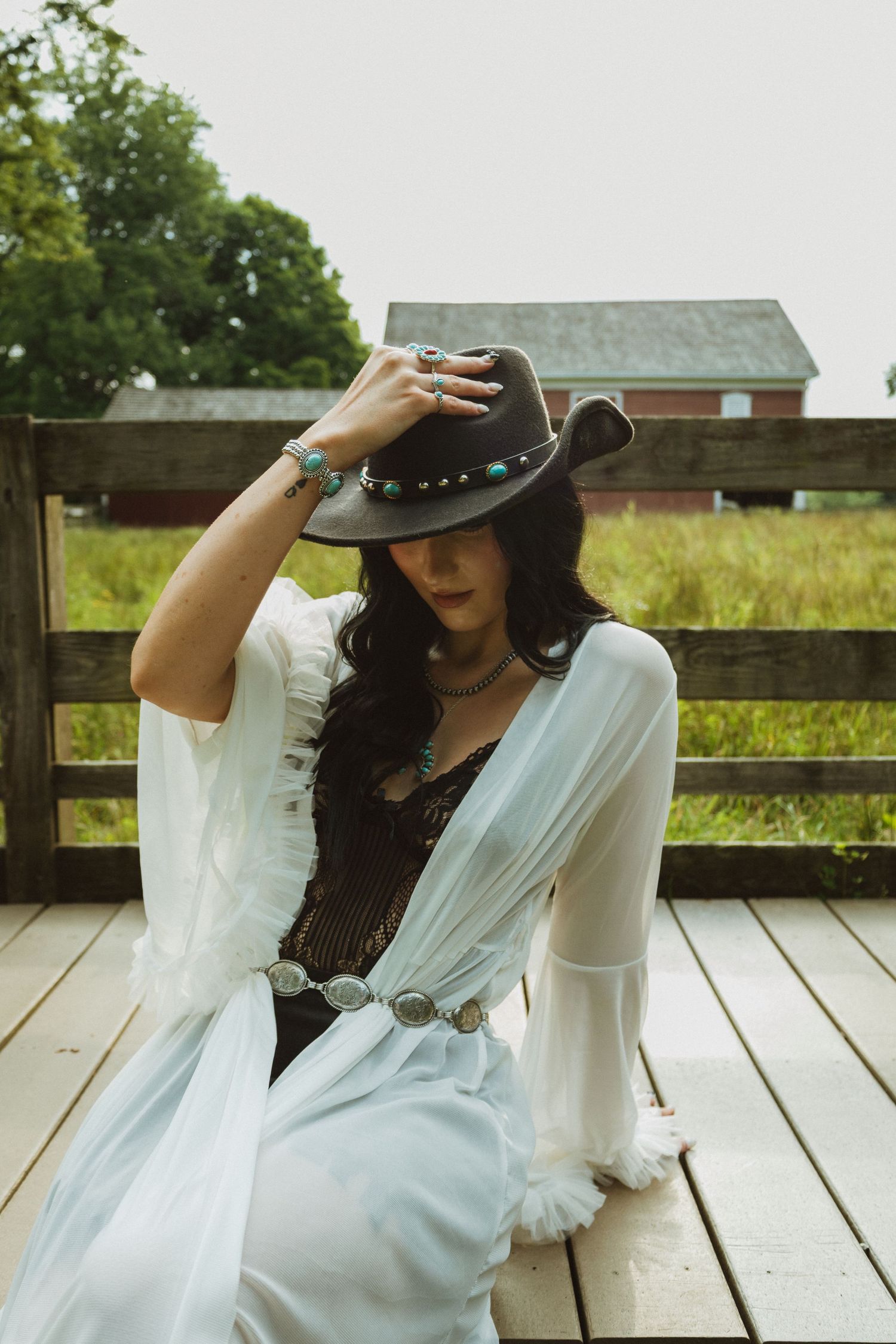 A woman in a flowing white dress and black cowboy hat sits on a wooden deck against a rustic barn backdrop.