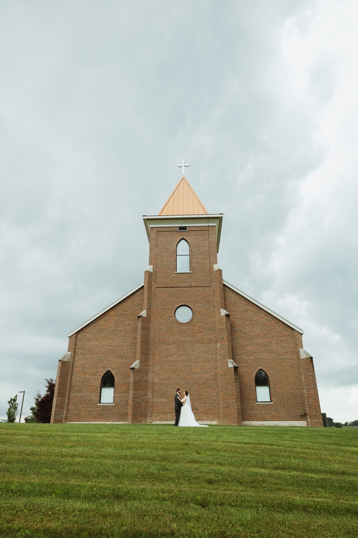 A brick church with a tall steeple sits atop a grassy hill against a cloudy sky.