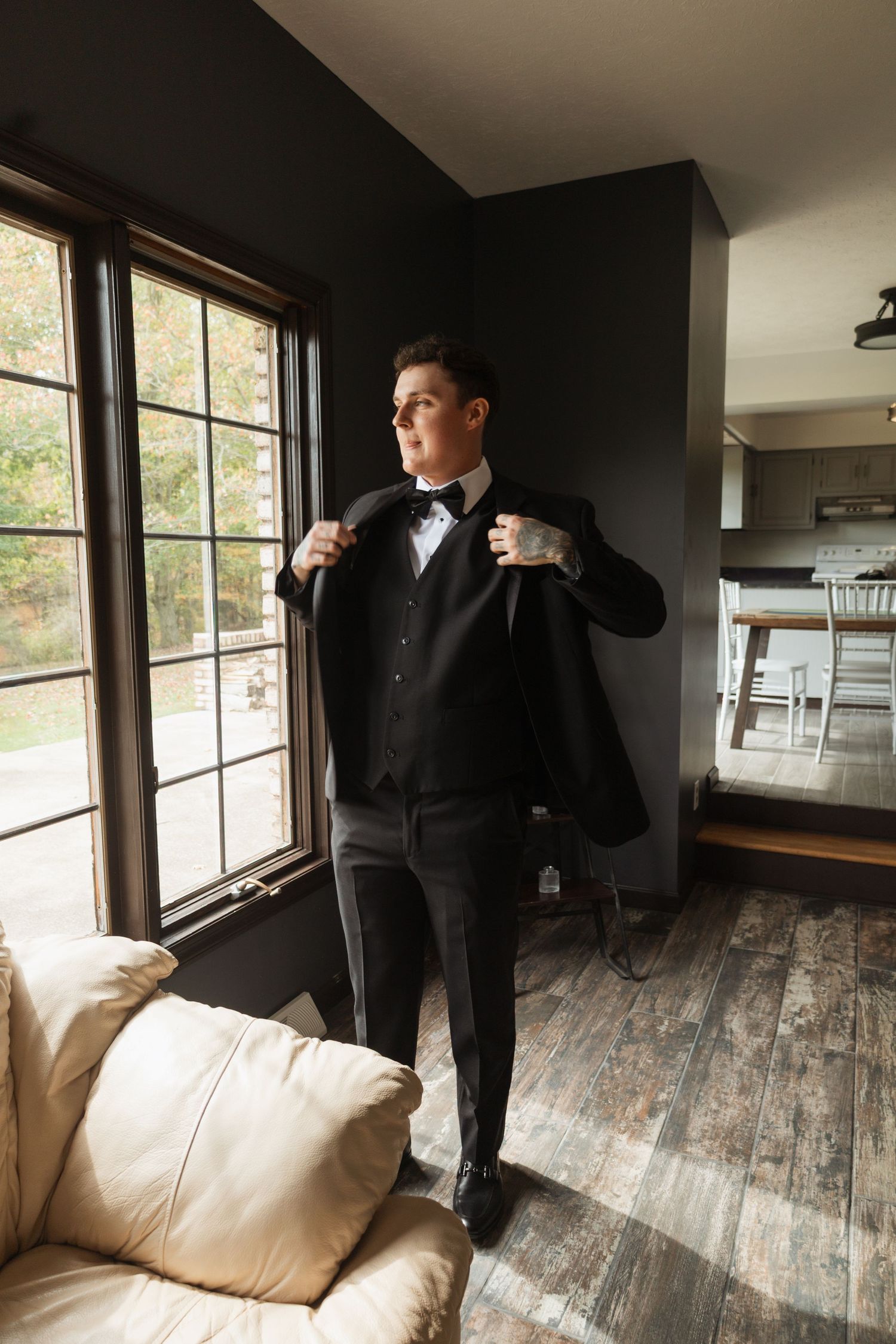 Person in formal black tuxedo adjusting outfit by window in modern interior.
