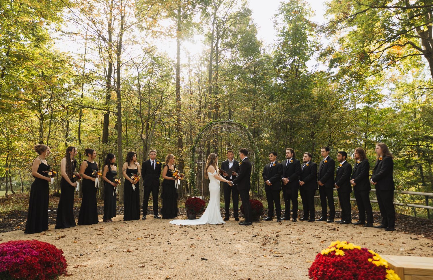 Wedding party lineup in a scenic outdoor setting with autumn trees and red floral arrangements.