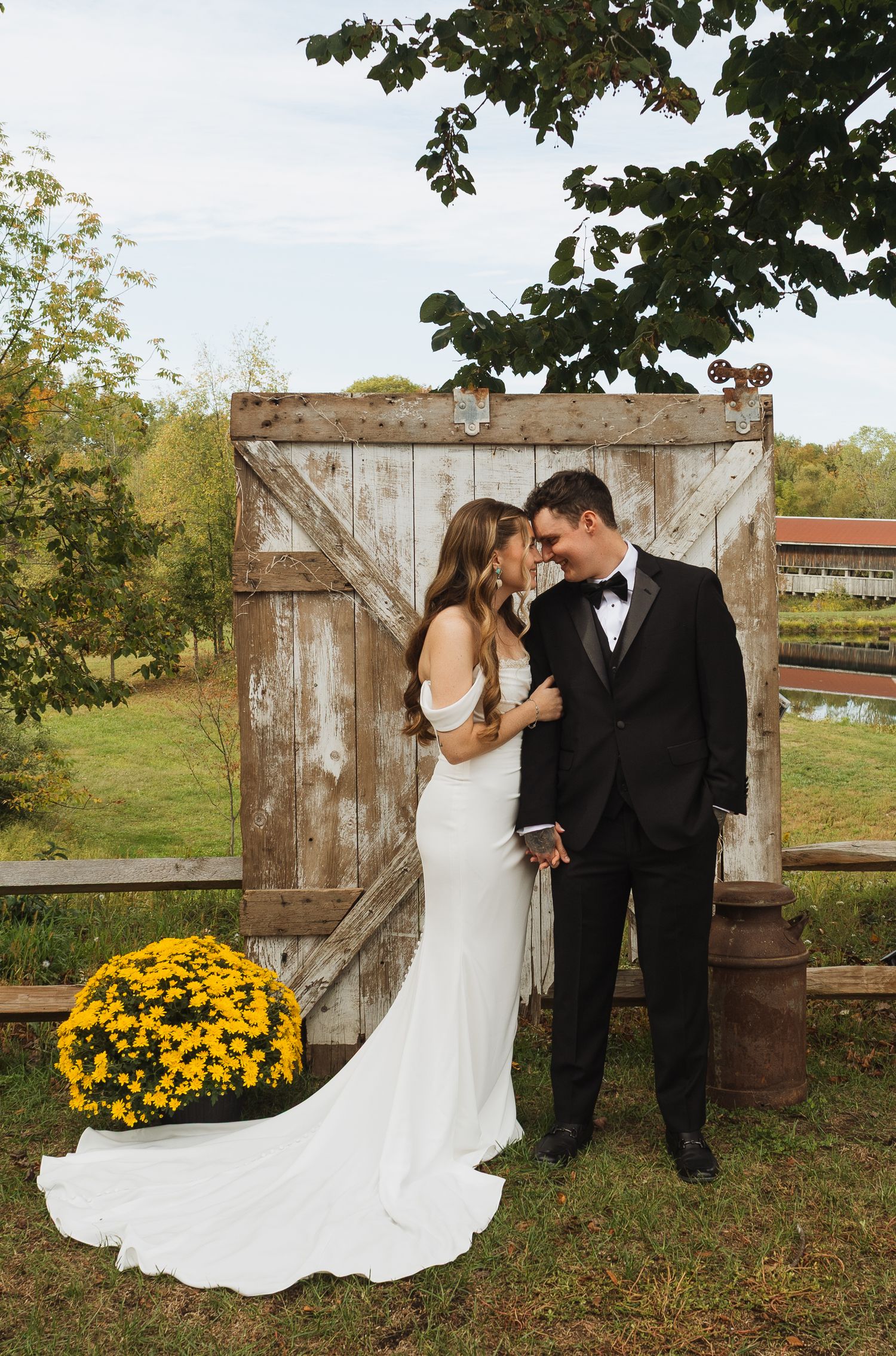 A romantic wedding couple poses in front of a rustic barn door with yellow mums decorating the autumn countryside scene.