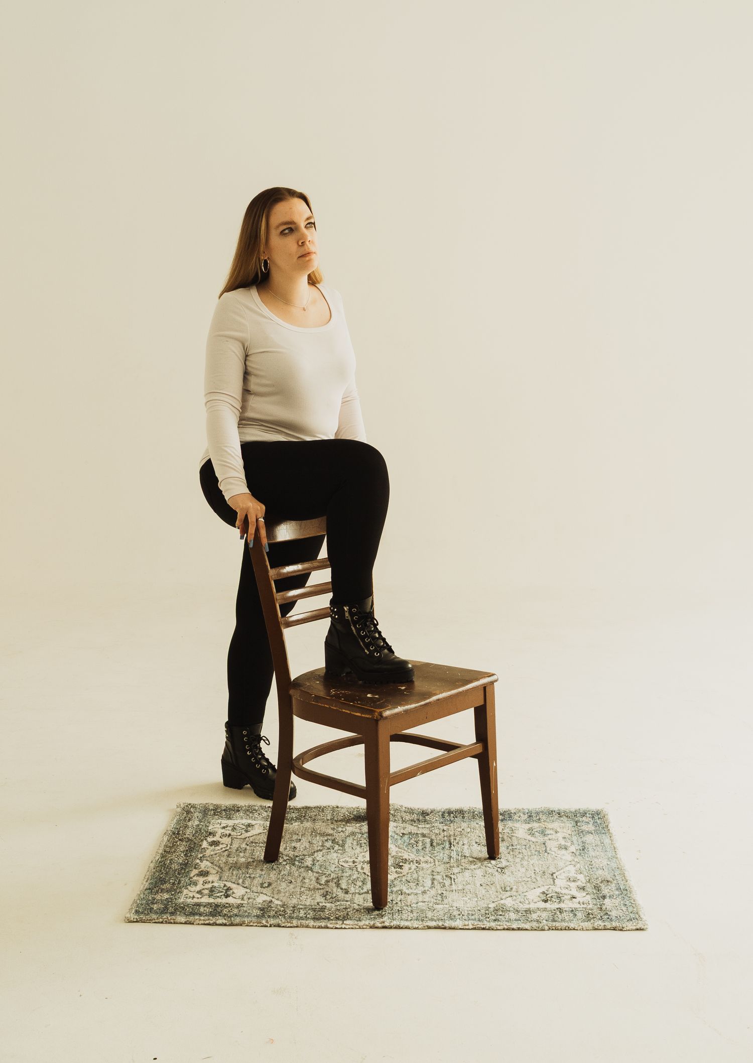 Person in white sweater and black pants striking a pose while standing on wooden chair on patterned rug.