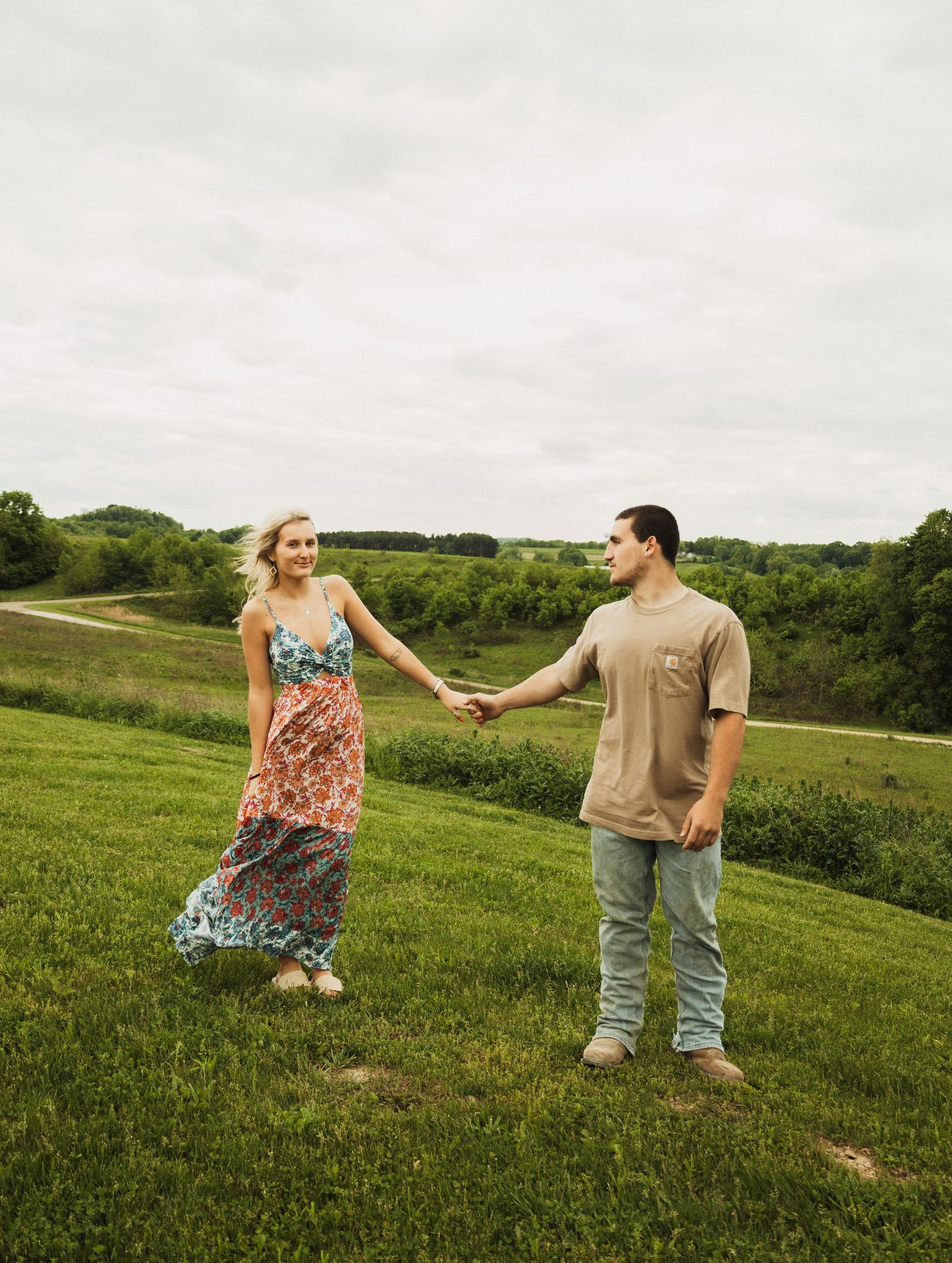 A couple stands hand-in-hand on a scenic grassy hill with a cloudy sky and verdant landscape behind them.