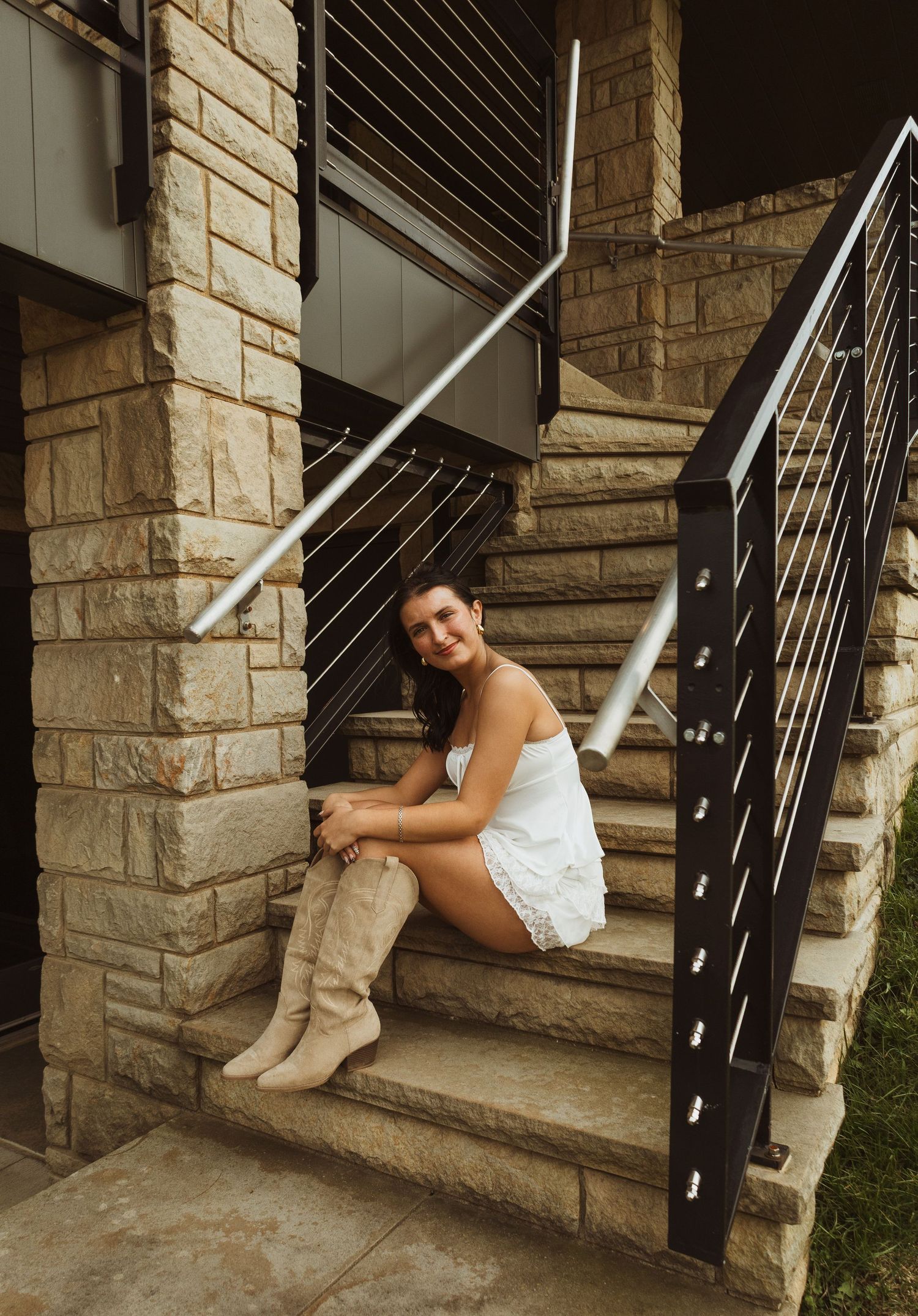Woman in white romper and tan knee-high boots sitting on stone steps with modern metal railing.