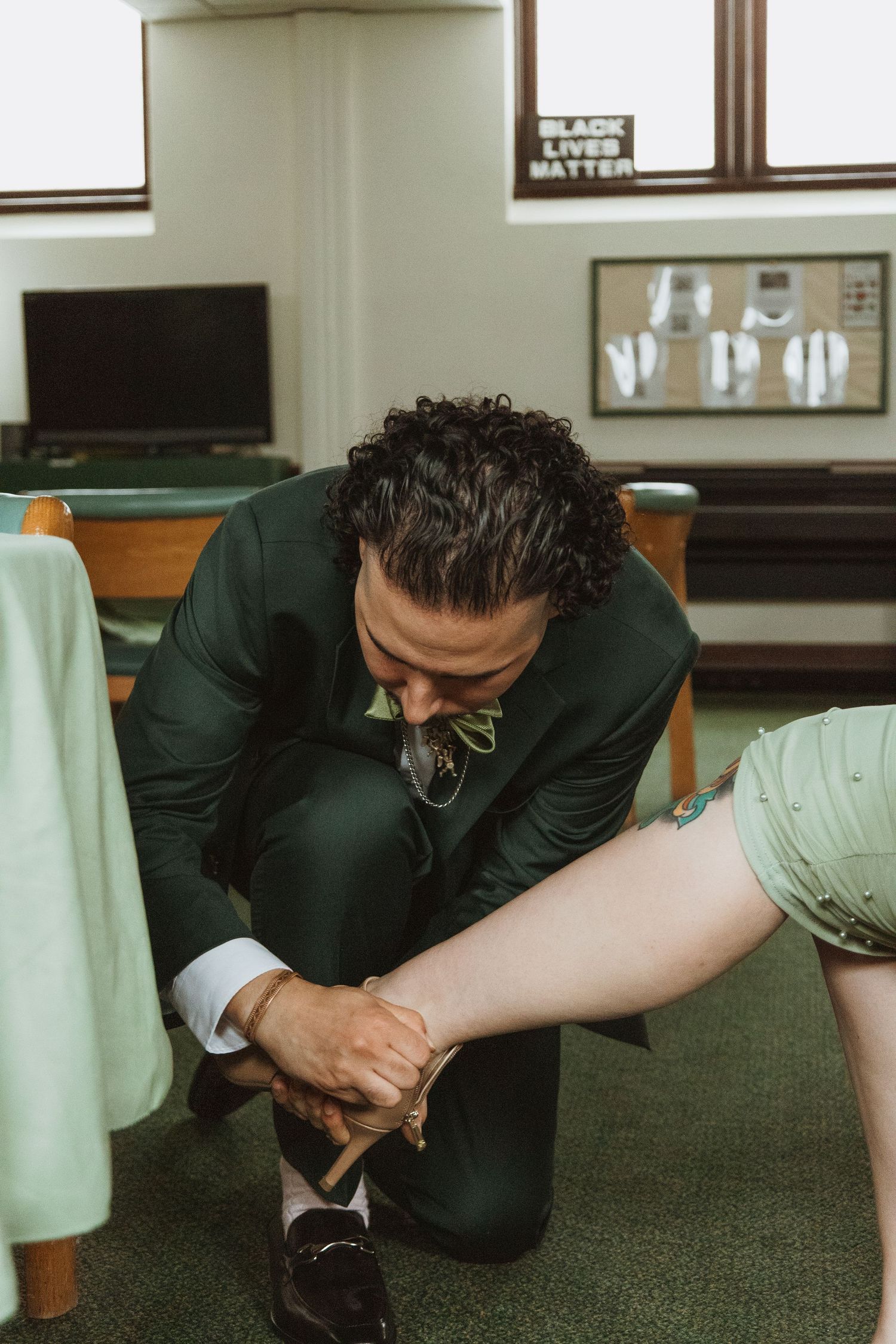 Groom in green suit kneeling to fasten bride's ankle strap shoe in a carpeted church room.