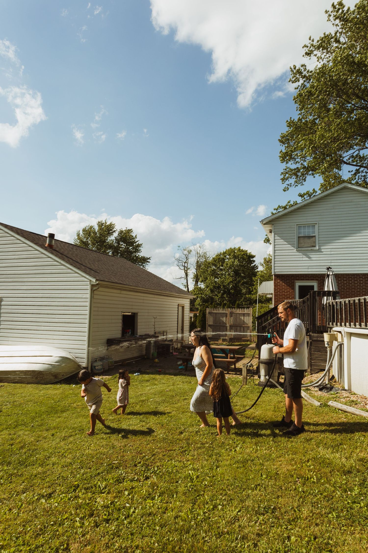 Family enjoying a sunny backyard day, with children playing on green grass beside two white suburban homes.