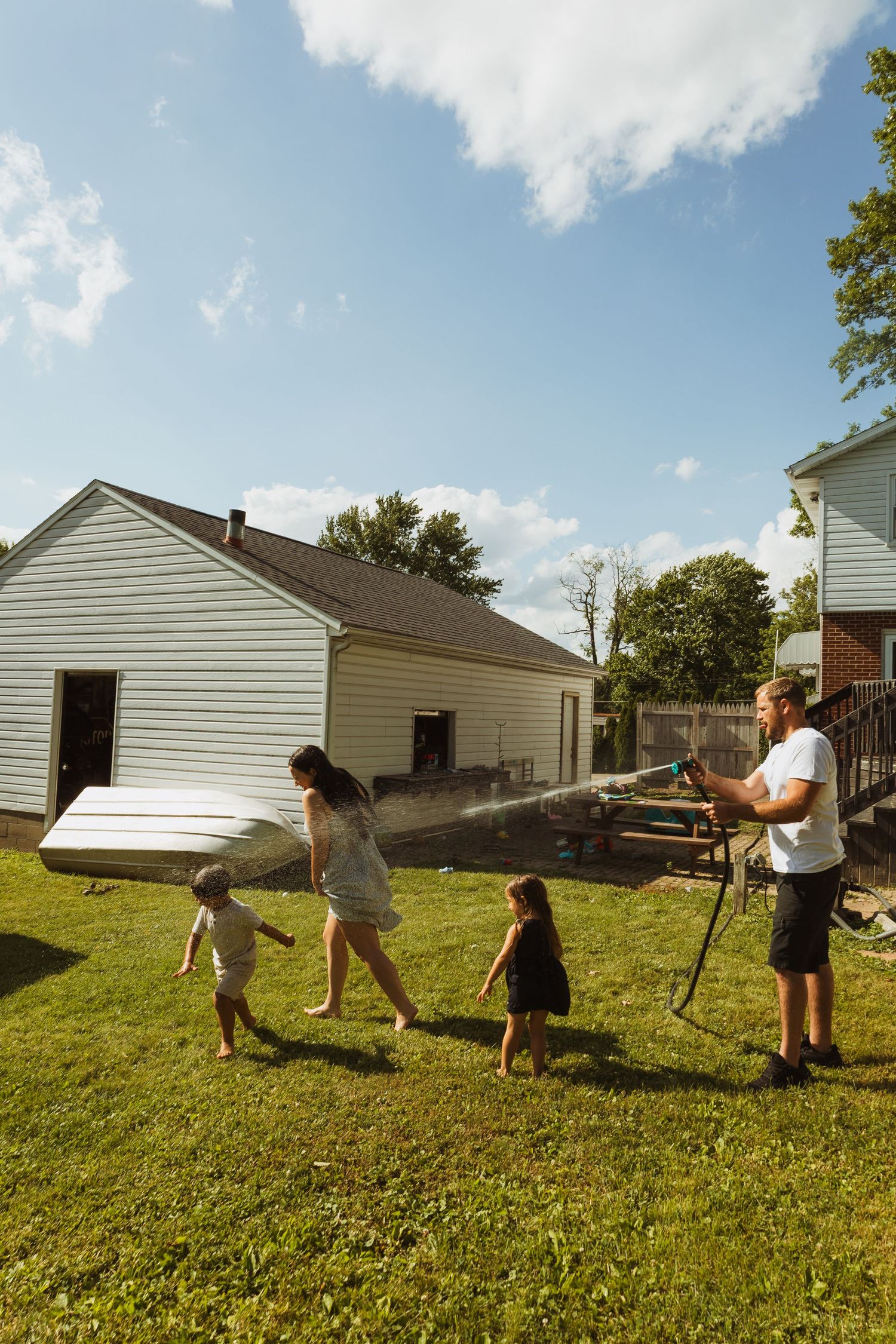 Family enjoying a sunny backyard day, with man spraying water hose while woman walks with two young children on green grass.
