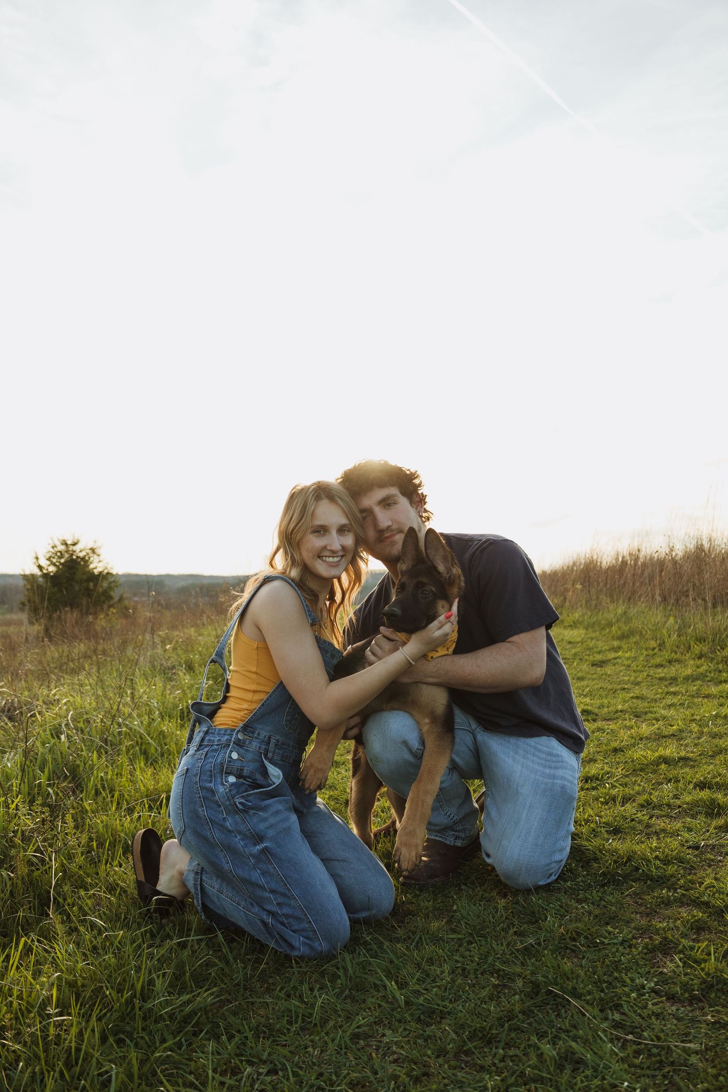 Smiling couple kneeling on green grass at sunset, holding a small dark dog in a scenic outdoor field.
