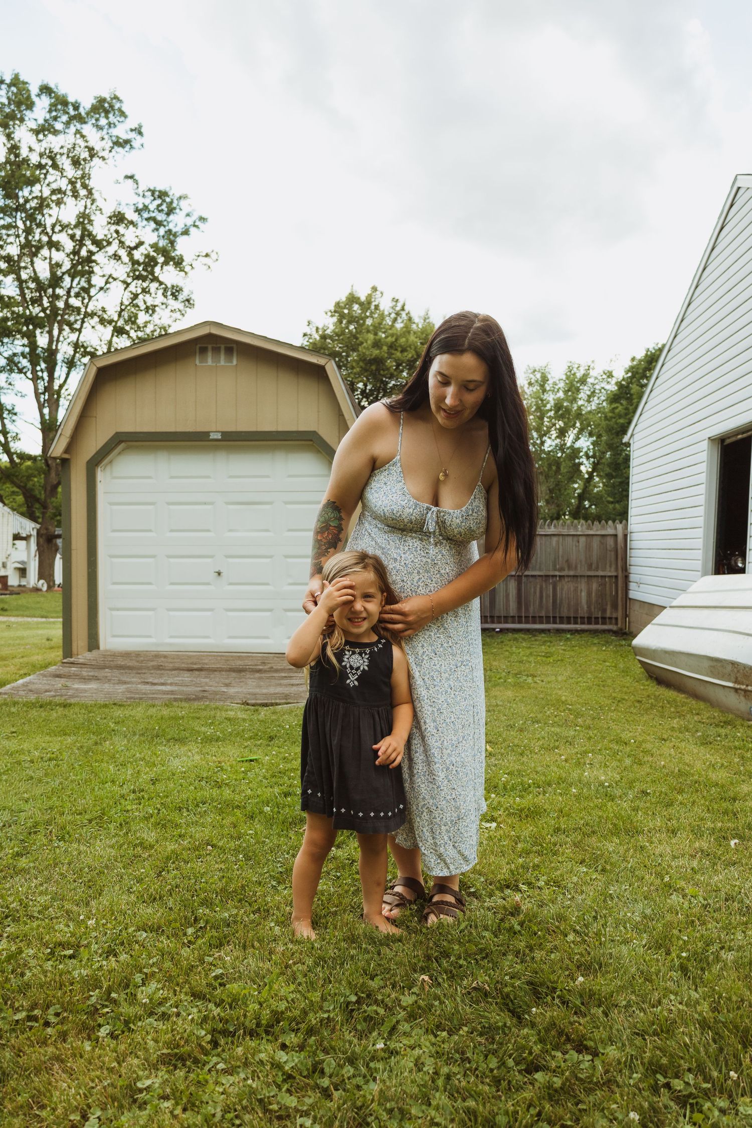 A young mother in a floral dress stands with her little girl in a backyard, gently touching her face near a garage.