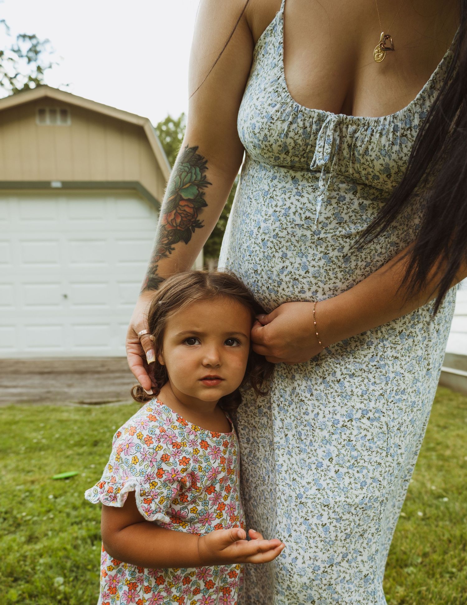 A tattooed pregnant woman in a floral dress stands outdoors with a young toddler girl leaning against her.