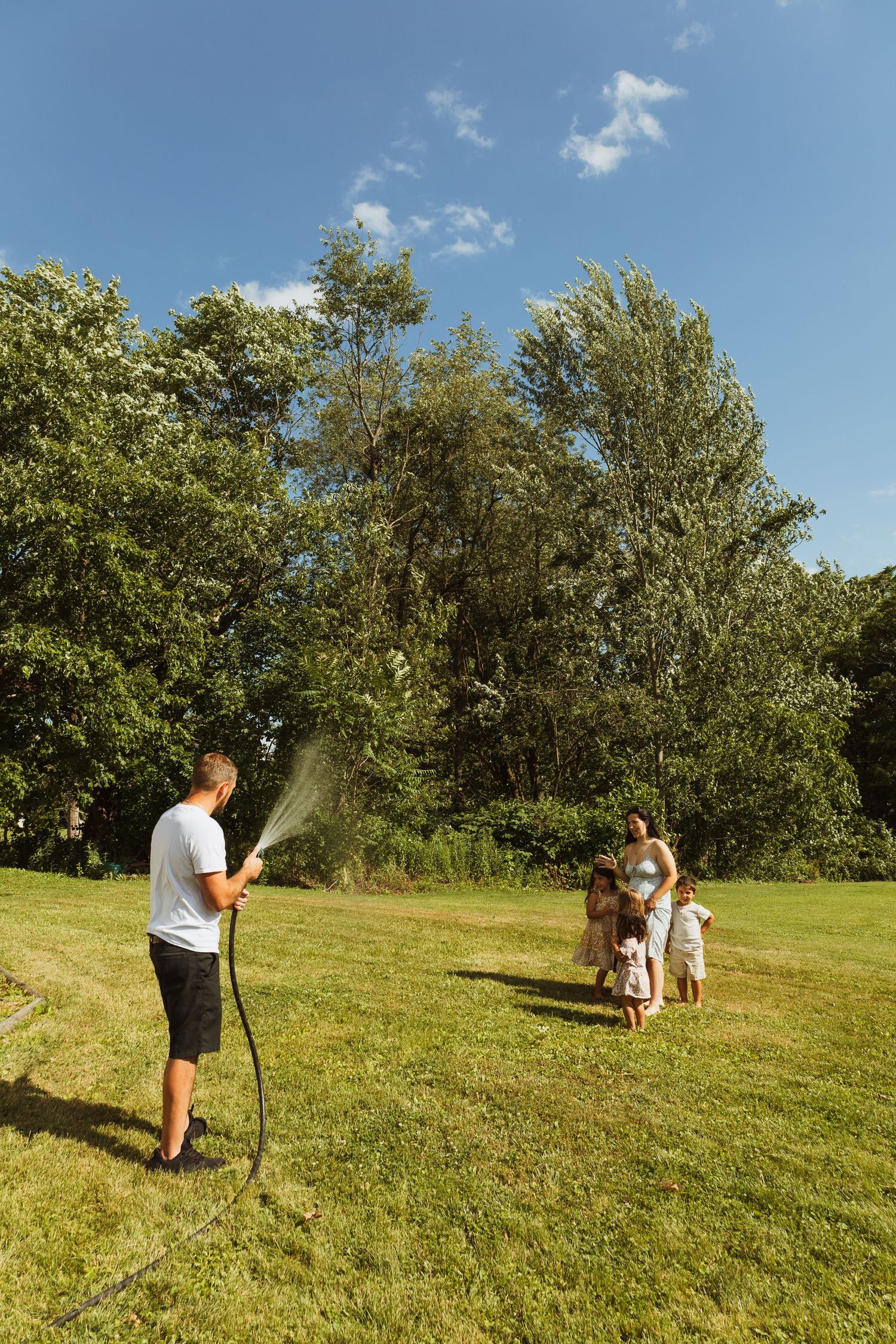 Family enjoying summer day outdoors, man spraying water with garden hose on green lawn surrounded by trees.