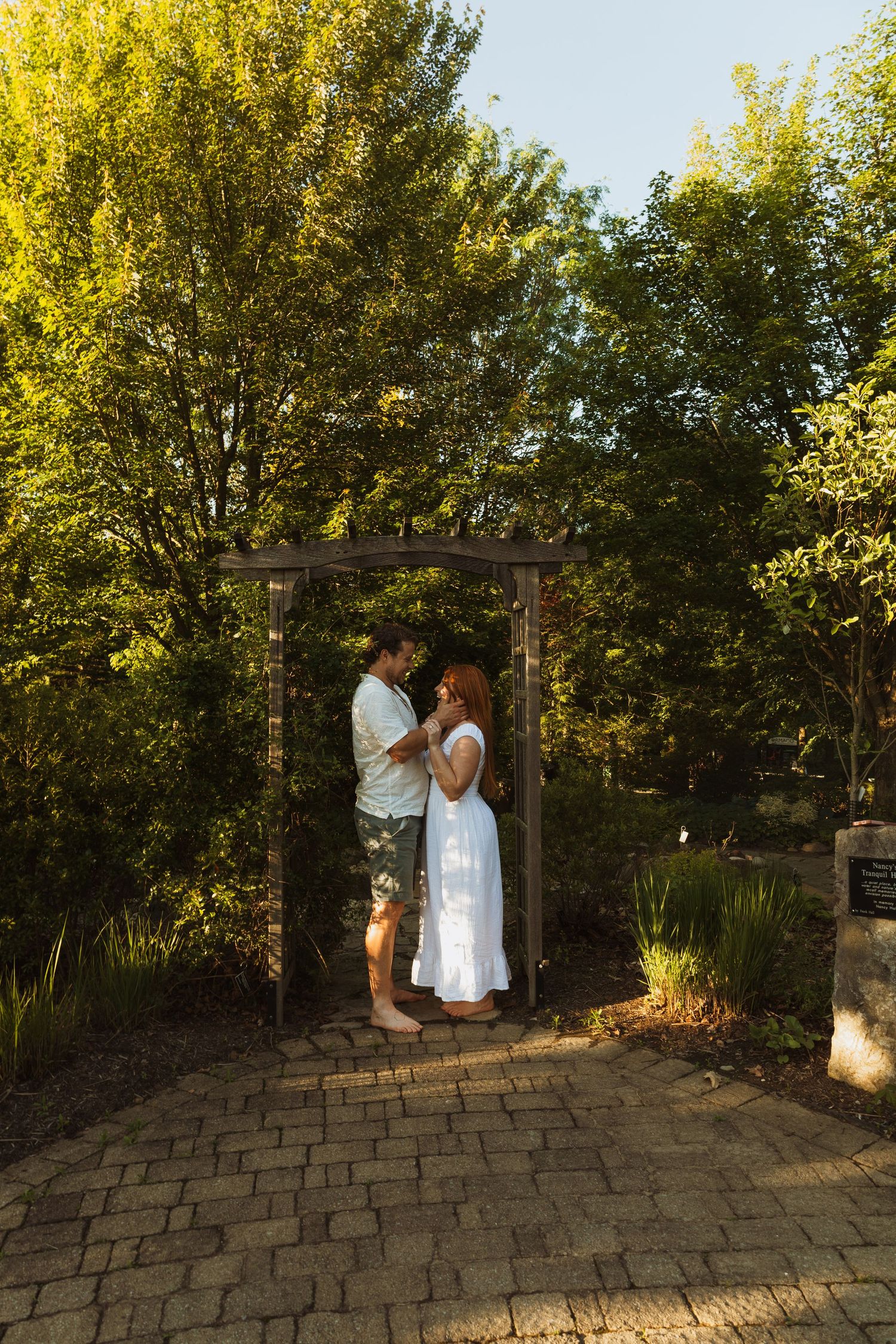 Couple embracing under a wooden garden arch surrounded by lush green trees on a sunny day.