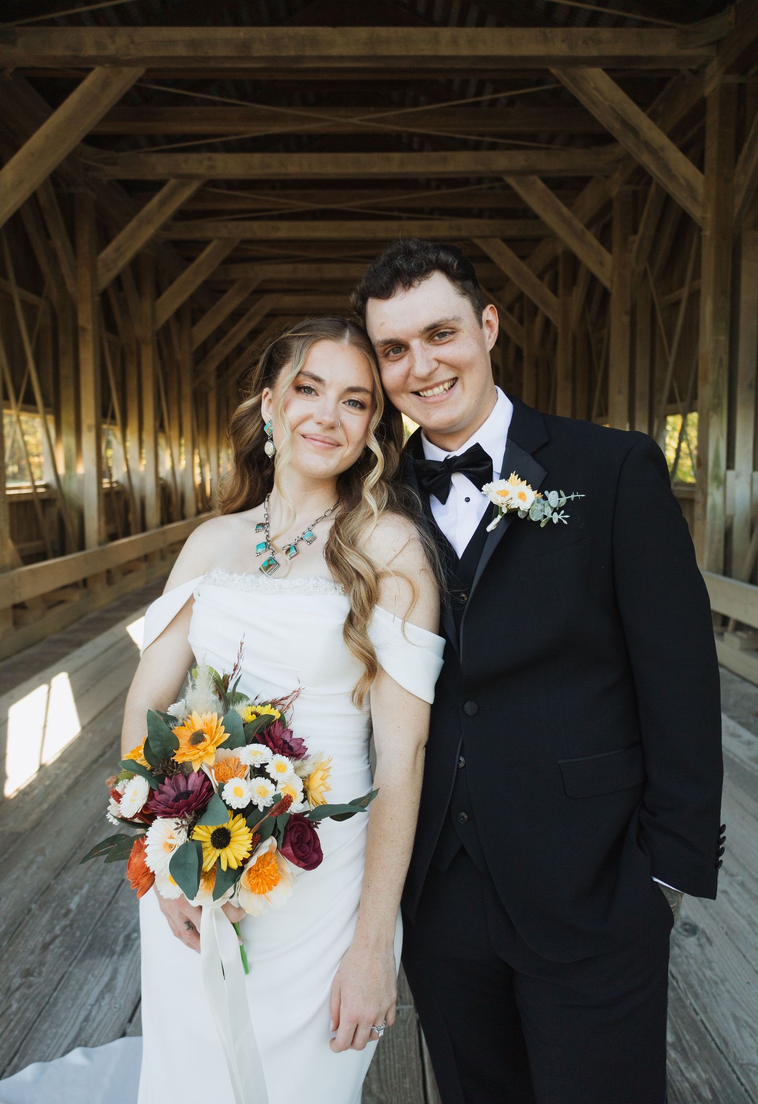 A romantic wedding couple poses together in a rustic covered wooden bridge with sunlight streaming through.