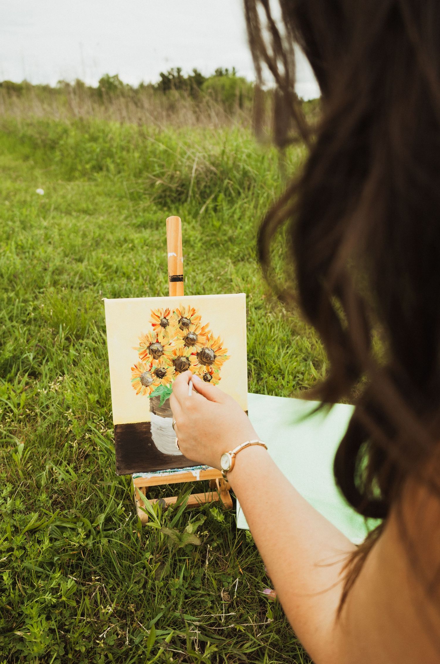 Artist painting yellow sunflowers in vase on small easel outdoors in grassy field during summer.