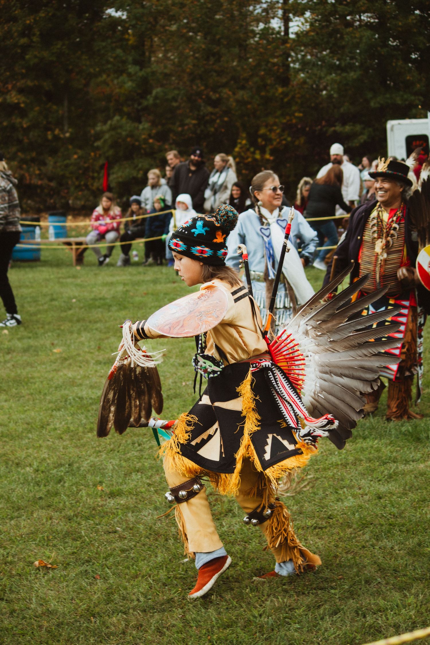 A dancer in traditional regalia with feather headdress performs energetic movements at an outdoor gathering.