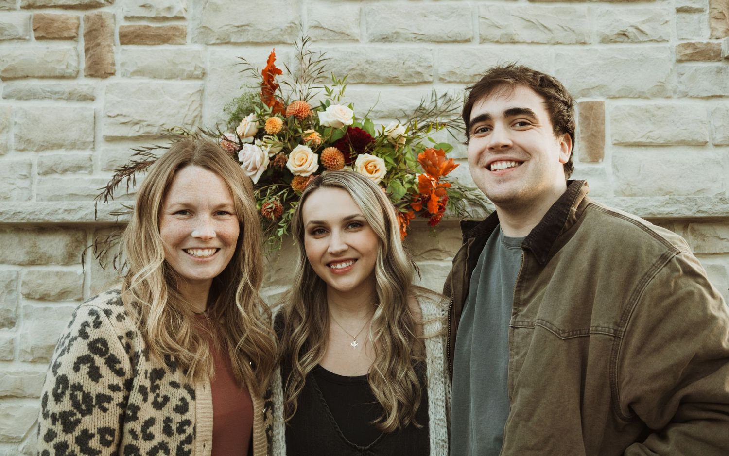 Three people pose together in front of stone wall with autumn floral arrangement.