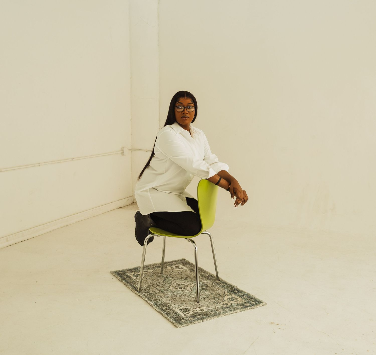 Person sits on a modern yellow and chrome chair on decorative rug against white wall in minimalist setting.