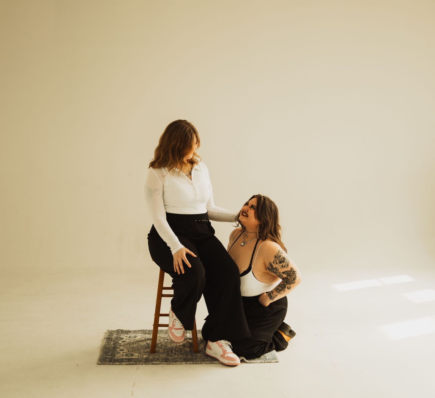 Two people in matching white tops and black pants posing with chairs in minimalist studio setting.