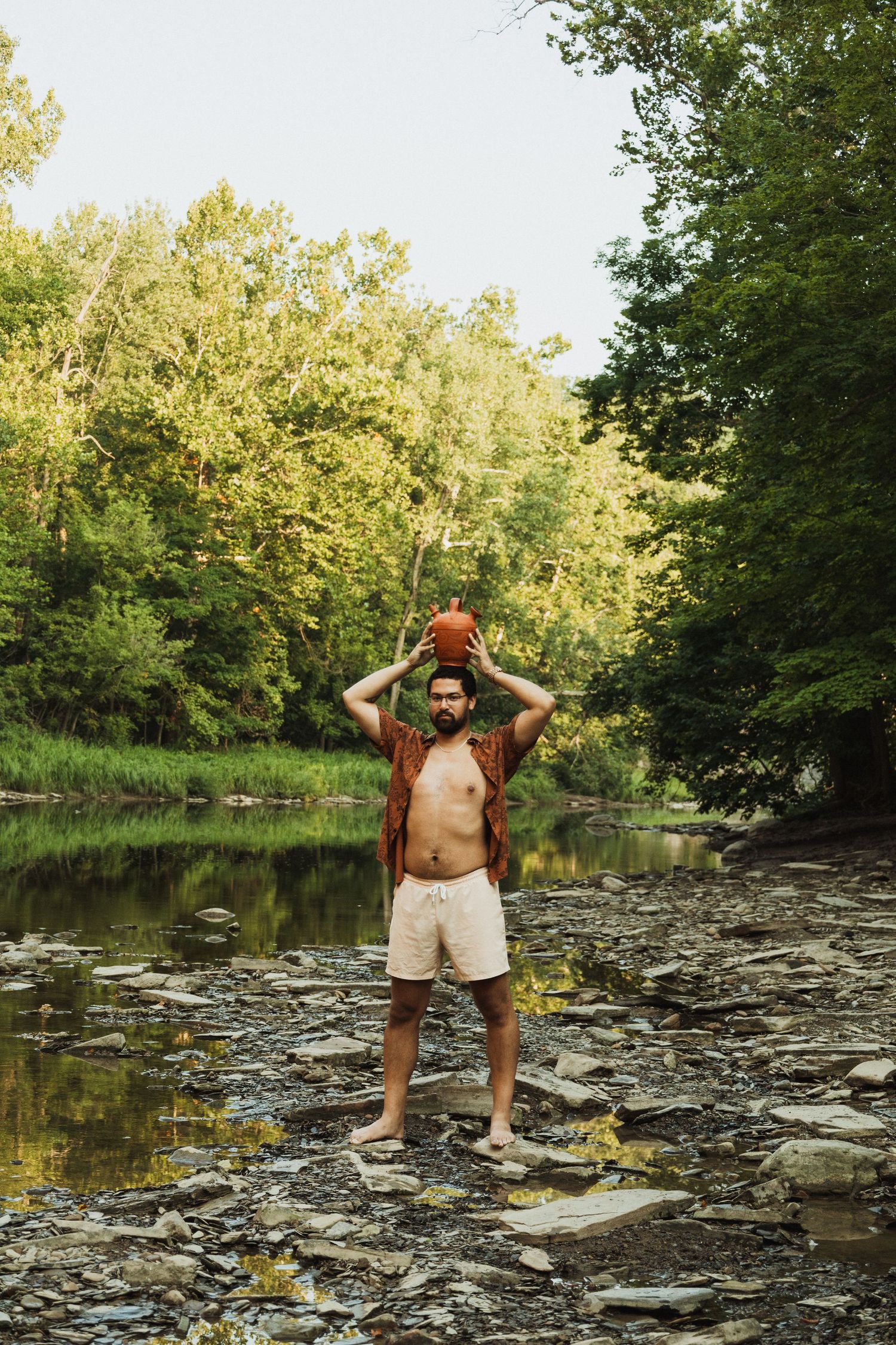 Person standing on rocky riverbed surrounded by lush green trees and still water.