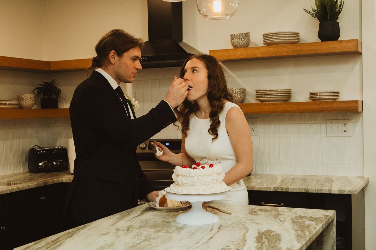 A playful cake-feeding moment between newlyweds in a modern kitchen setting.
