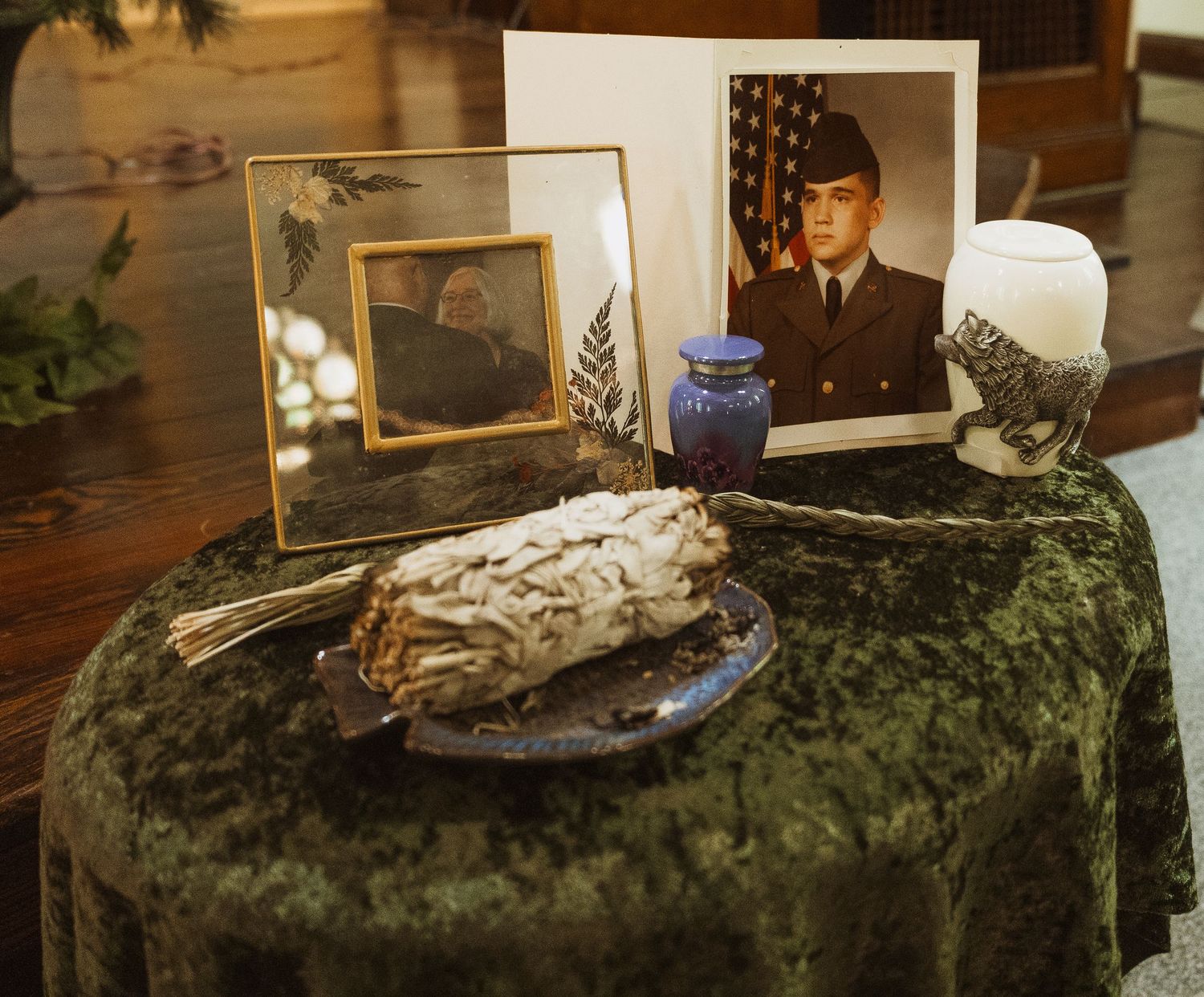 Memorial table with military portrait, framed photos, white sage bundle, blue urn, and candle on green velvet cloth.