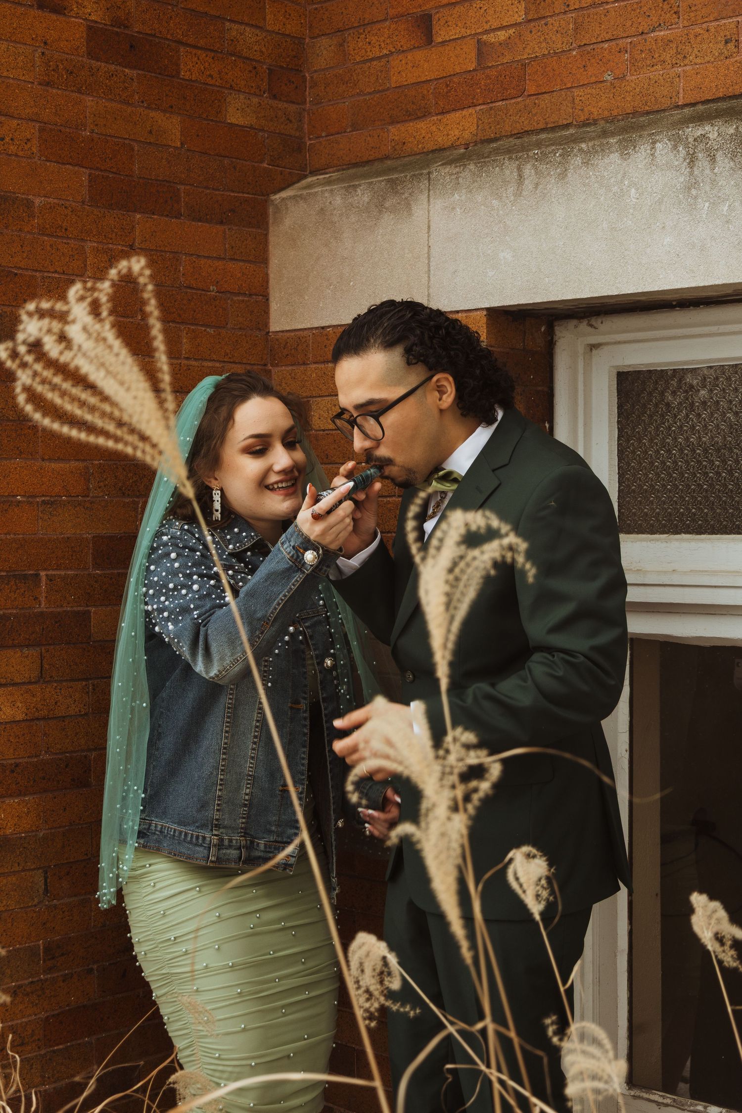 Bride in green wedding dress and veil applies lipstick to groom near brick wall with dried grasses.